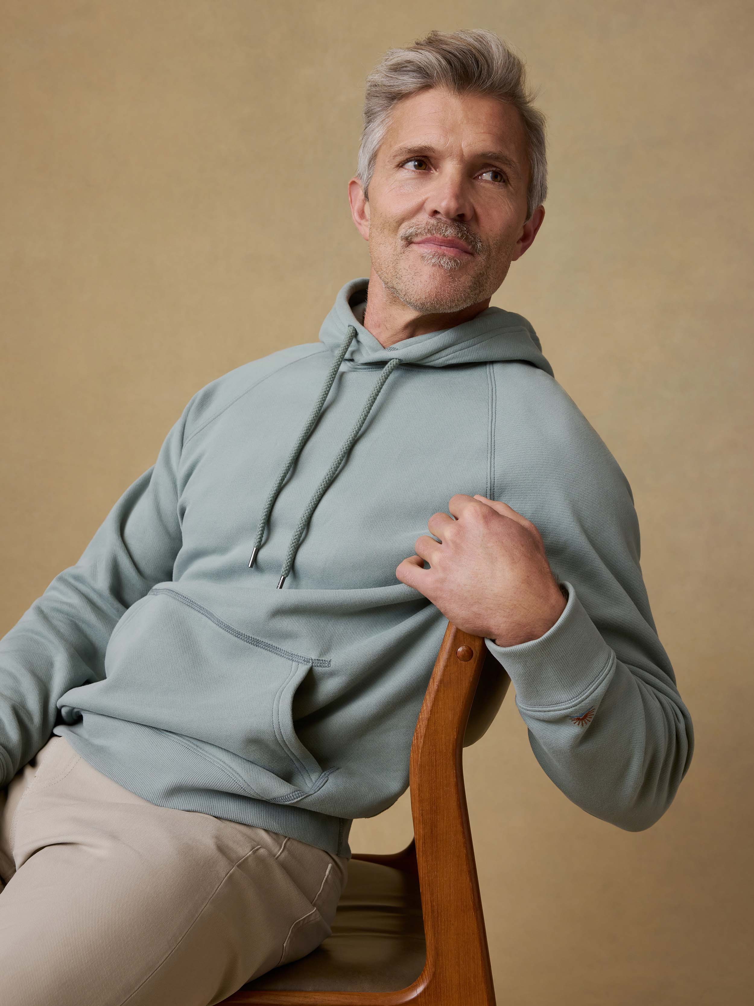 Man is sitting on a wooden chair wearing a light green hoodie and beige pants against a beige background