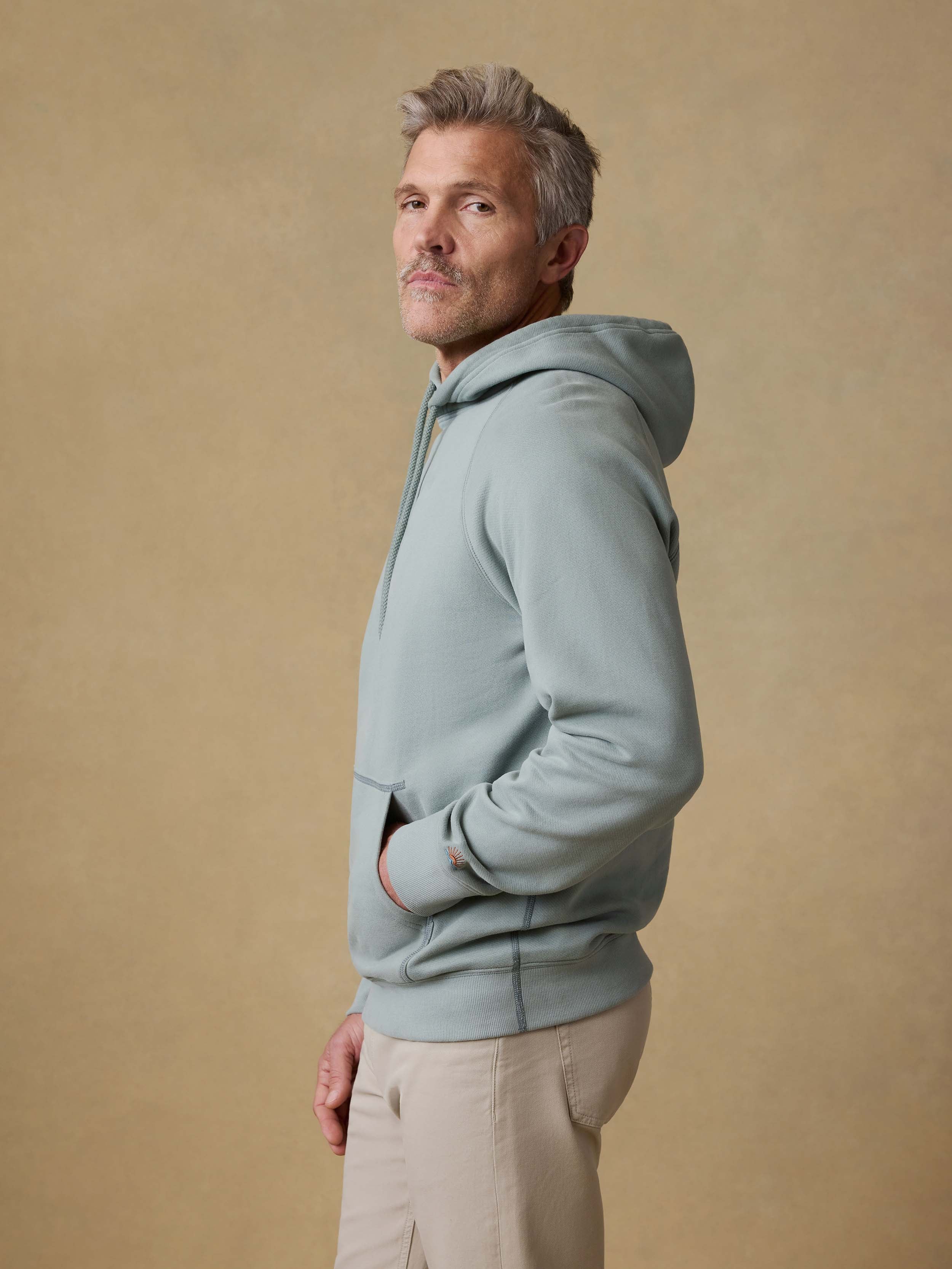 Man is wearing a light green hoodie and beige pants against a beige background