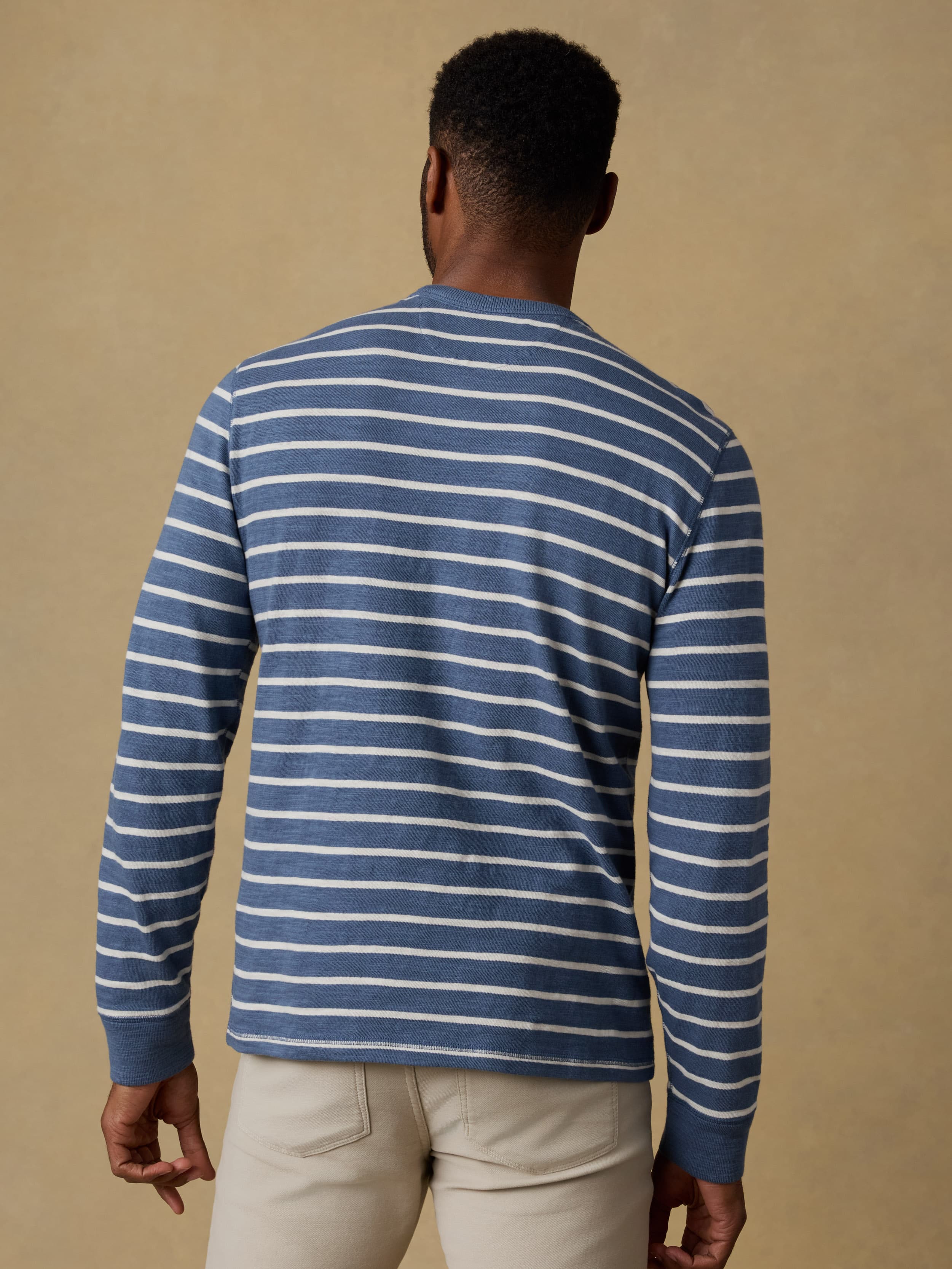 Back view of a man wearing a blue and white striped crewneck sweatshirt and beige pants against a beige wall.