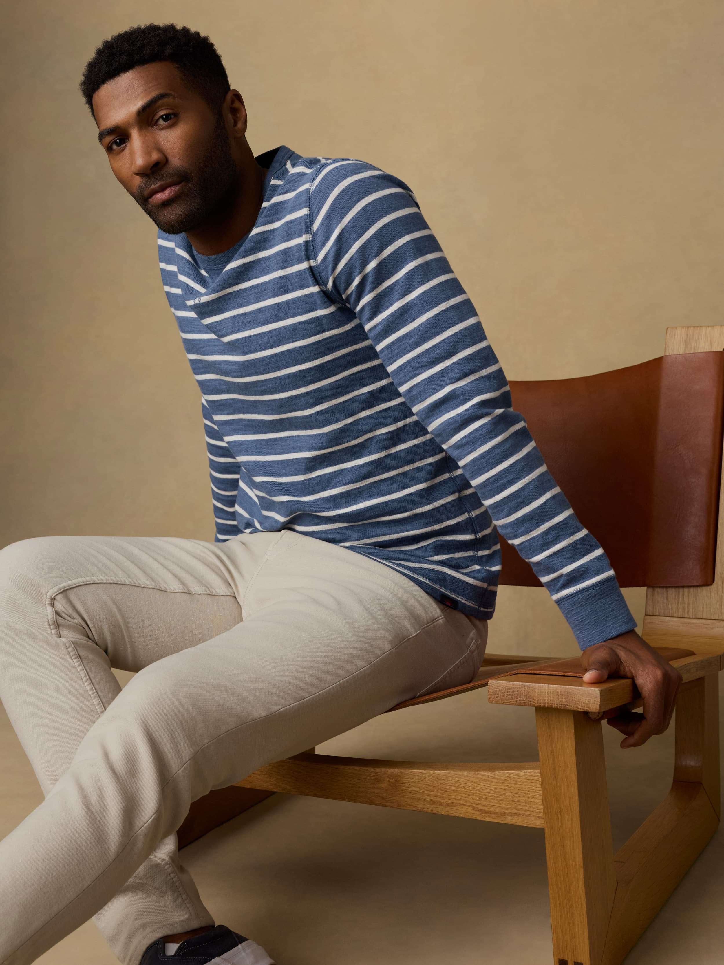 Man sitting on a wooden chair wearing a blue and white striped crewneck sweatshirt and beige pants against a beige wall.