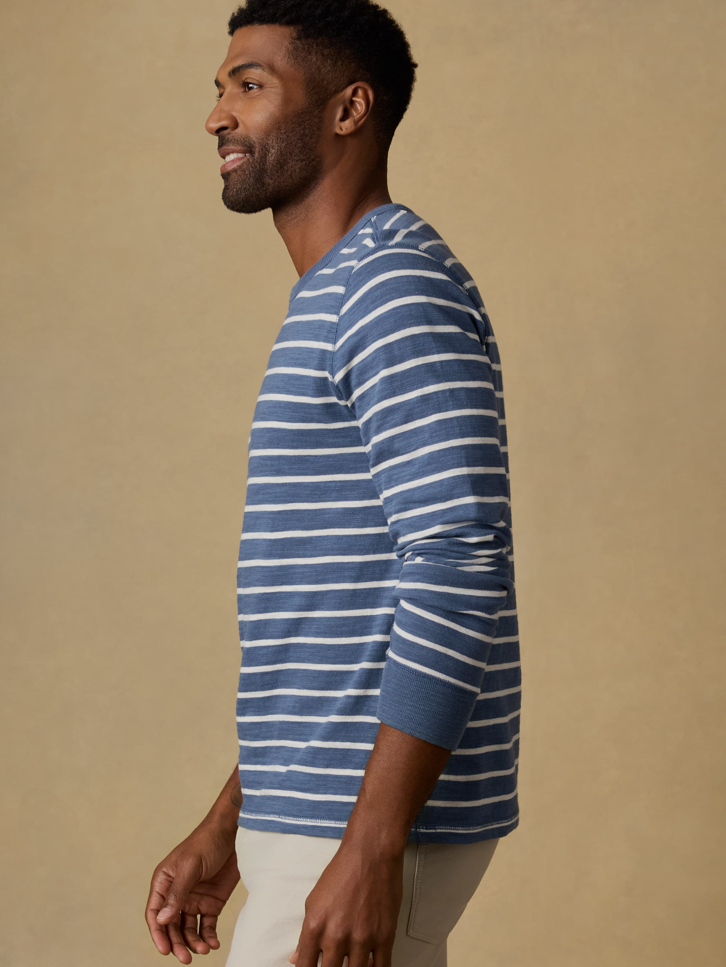 Man wearing a blue and white striped crewneck sweatshirt and beige pants against a beige wall.