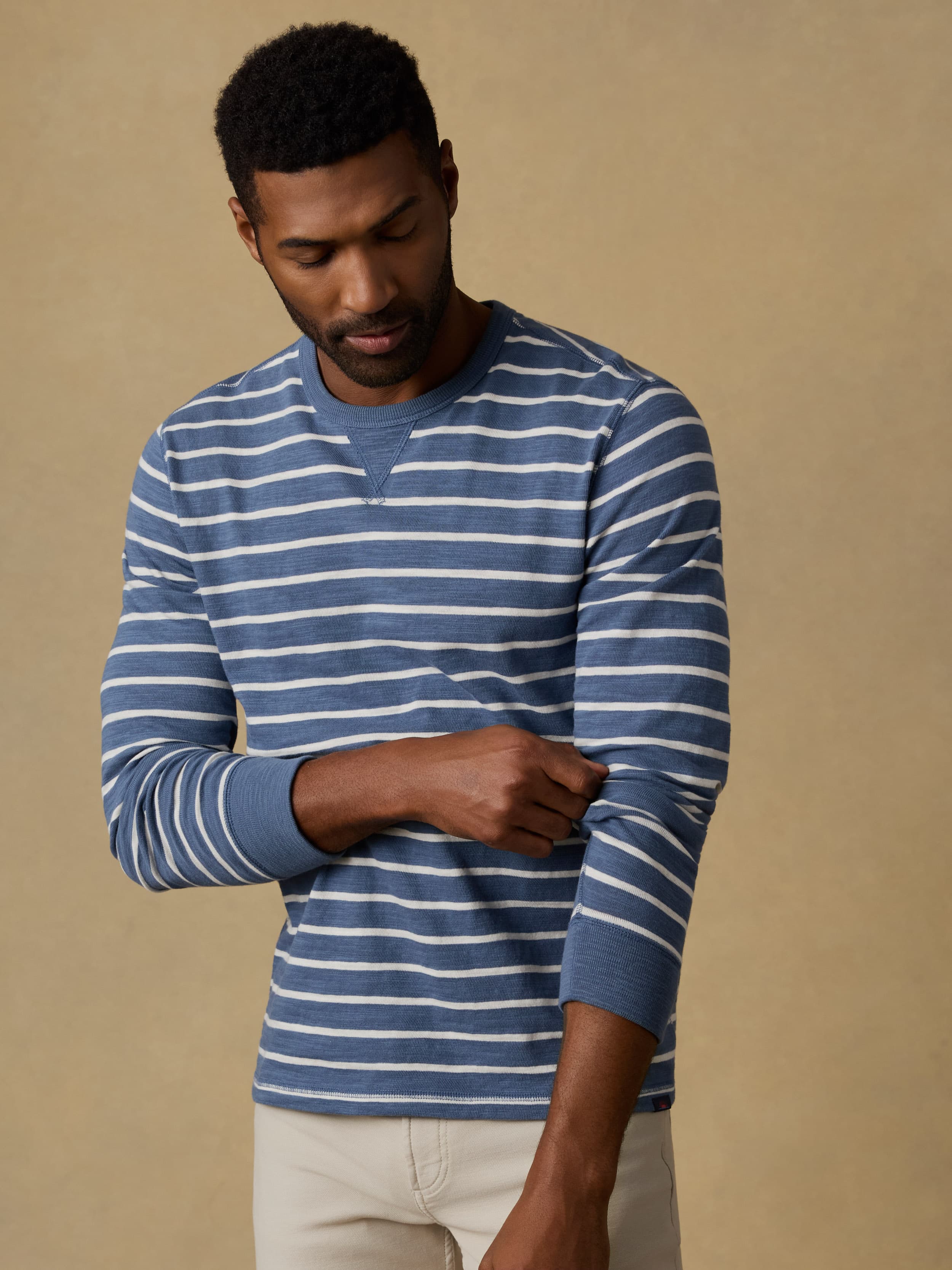 Man wearing a blue and white striped crewneck sweatshirt and beige pants against a beige wall.