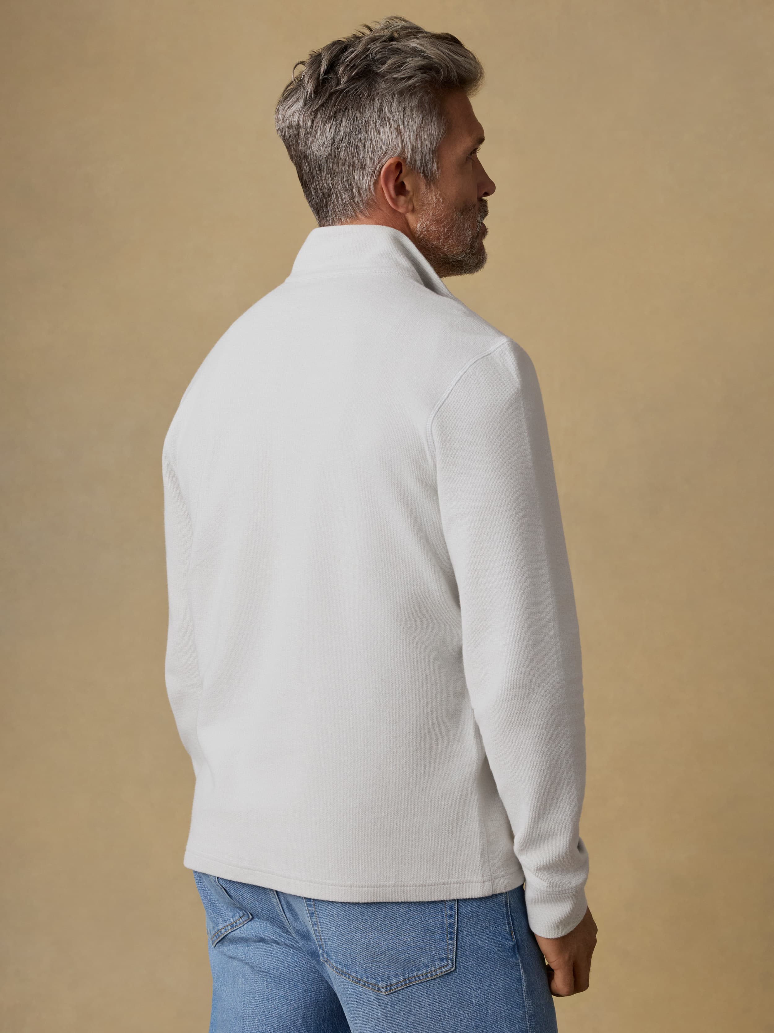 Back view of a man wearing a light gray quarter zip sweatshirt and blue jeans against a beige background