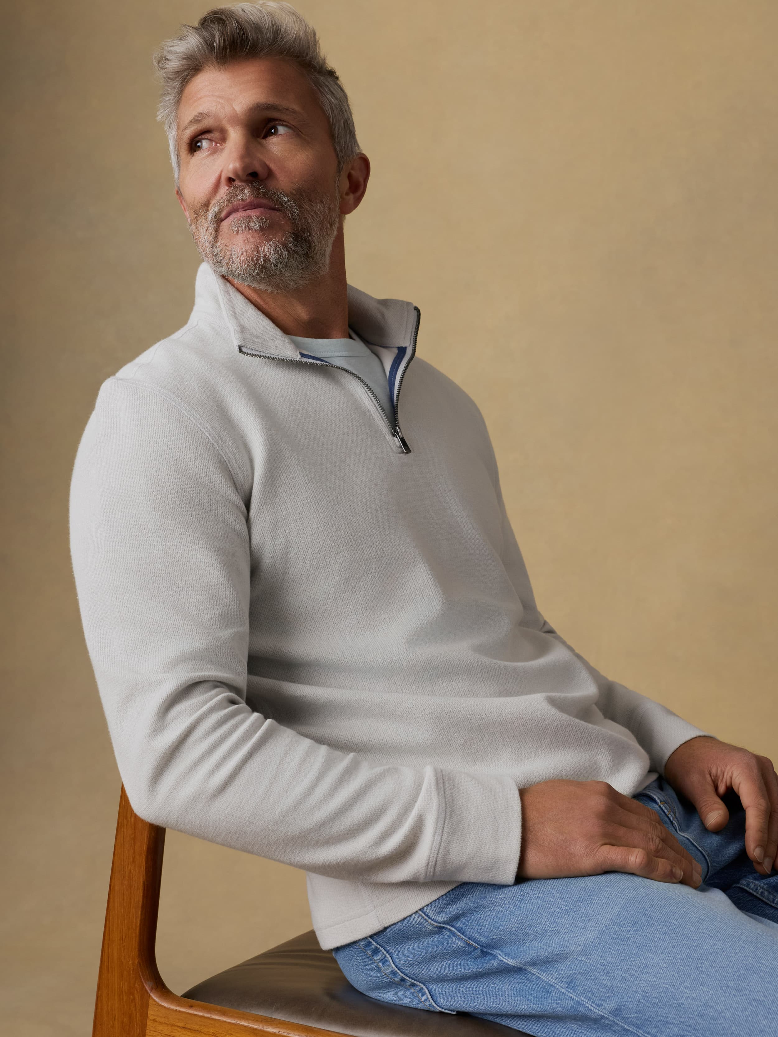 Man sitting on a wooden chair wearing a light gray quarter zip sweatshirt and blue jeans against a beige background