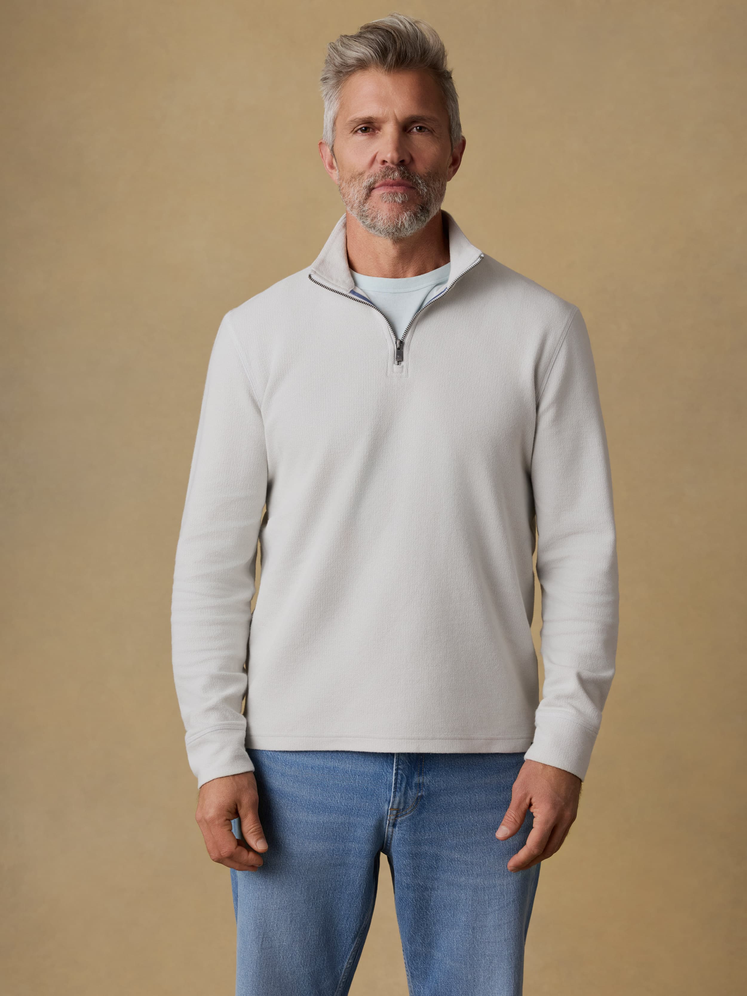 Man wearing a light gray quarter zip sweatshirt and blue jeans against a beige background