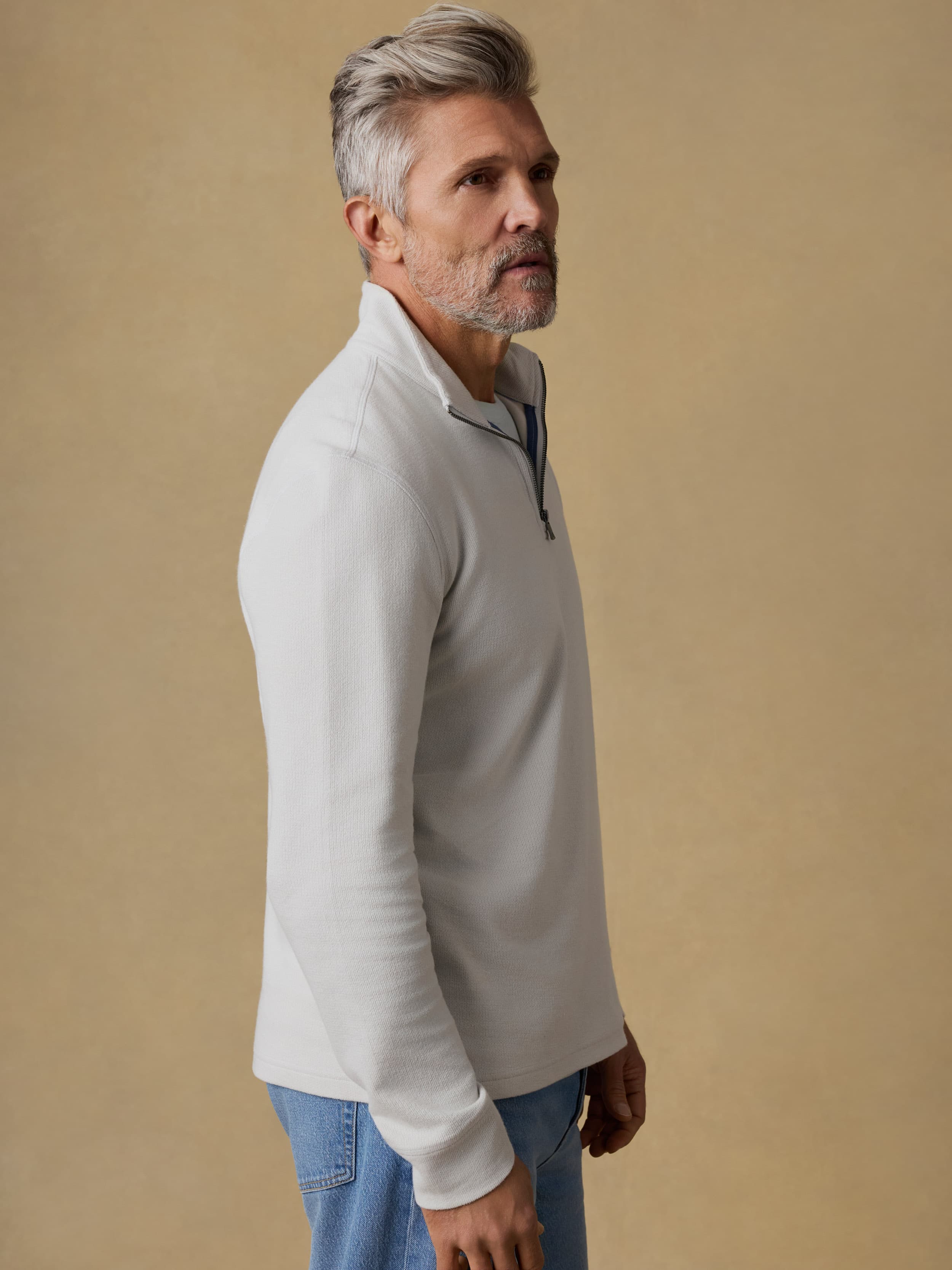 Man wearing a light gray quarter zip sweatshirt and blue jeans against a beige background