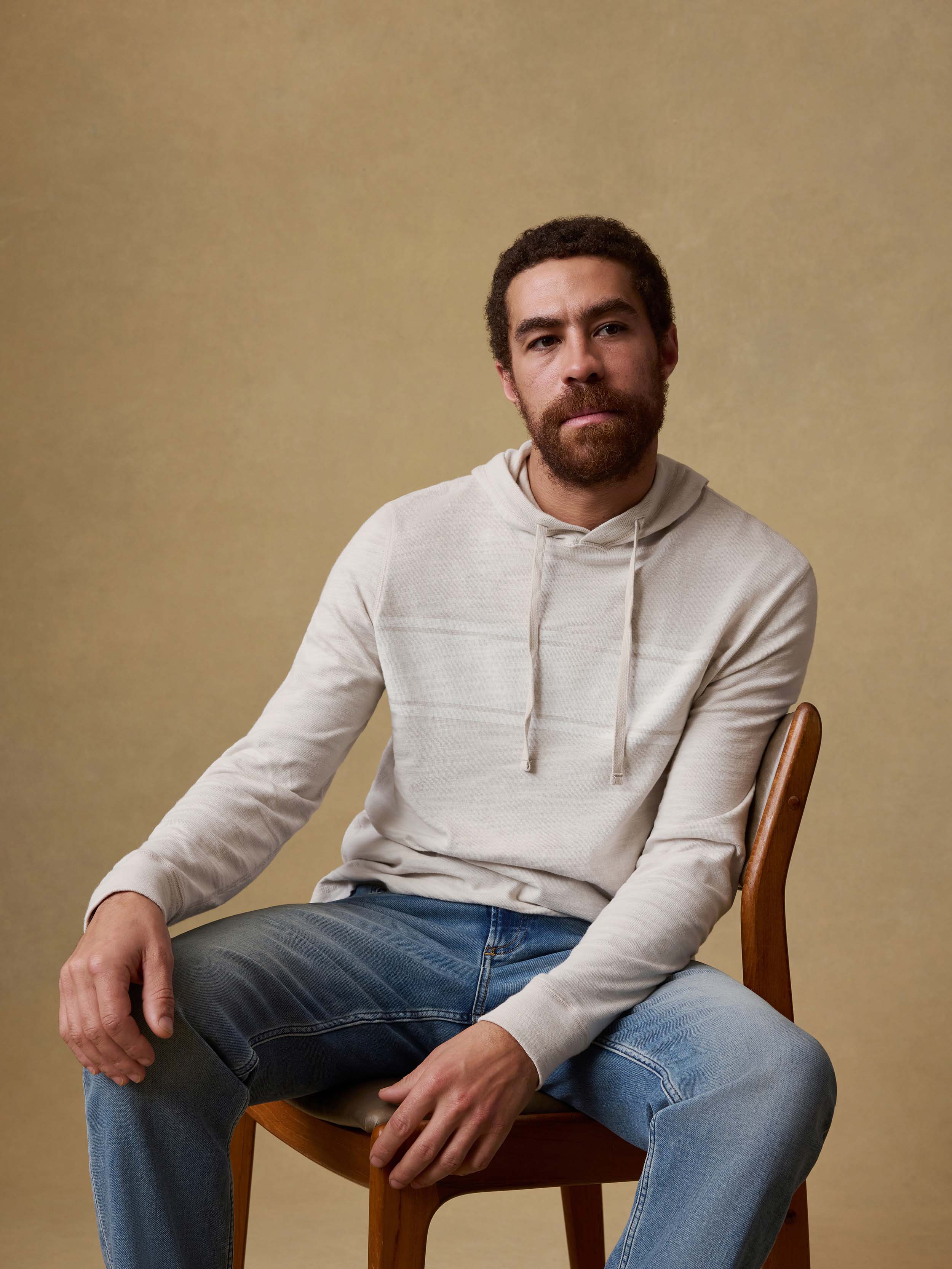 Man sitting on a wooden chair wearing a light gray hoodie and blue jeans against a beige background