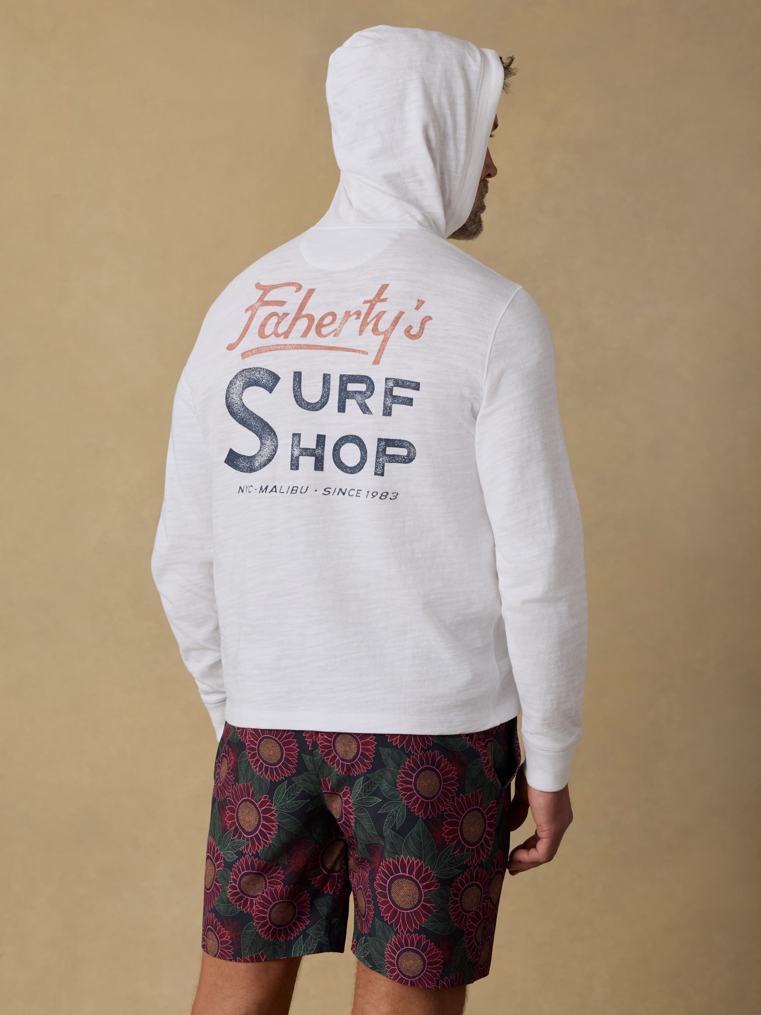 Back view of a man wearing a white hoodie with Faherty Surf Shop graphic on the back and patterned shorts on a beige background