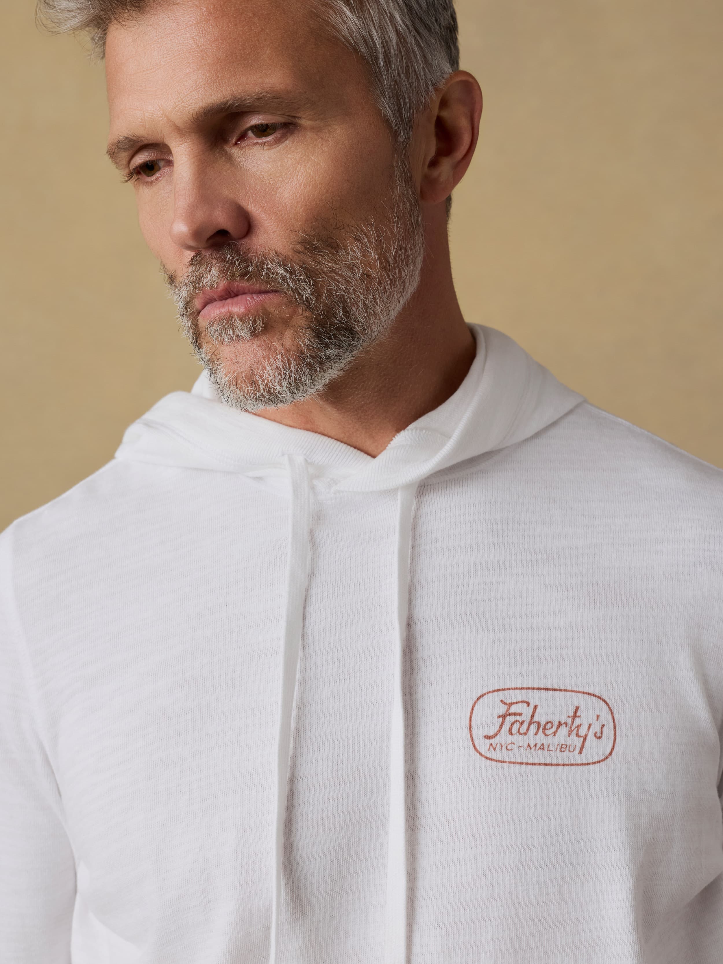 Man wearing a white hoodie with Faherty brand logo at the left chest and patterned shorts on a beige background