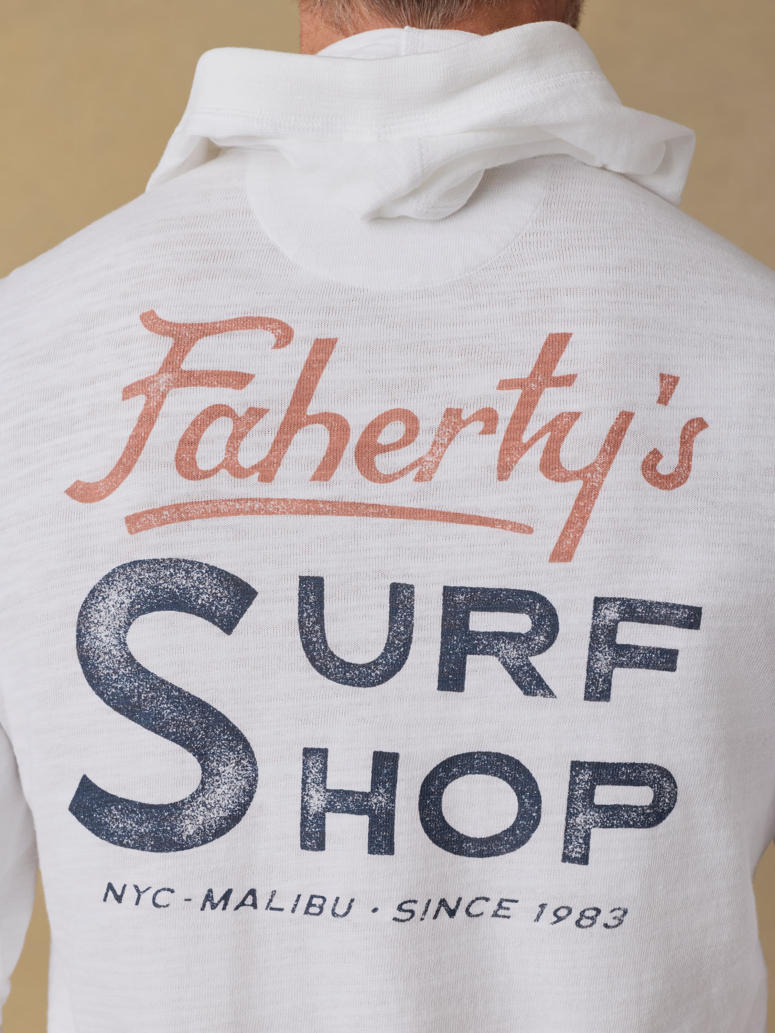 Back view of a man wearing a white hoodie with 'Faherty's Surf Shop' graphic on the back against a beige background