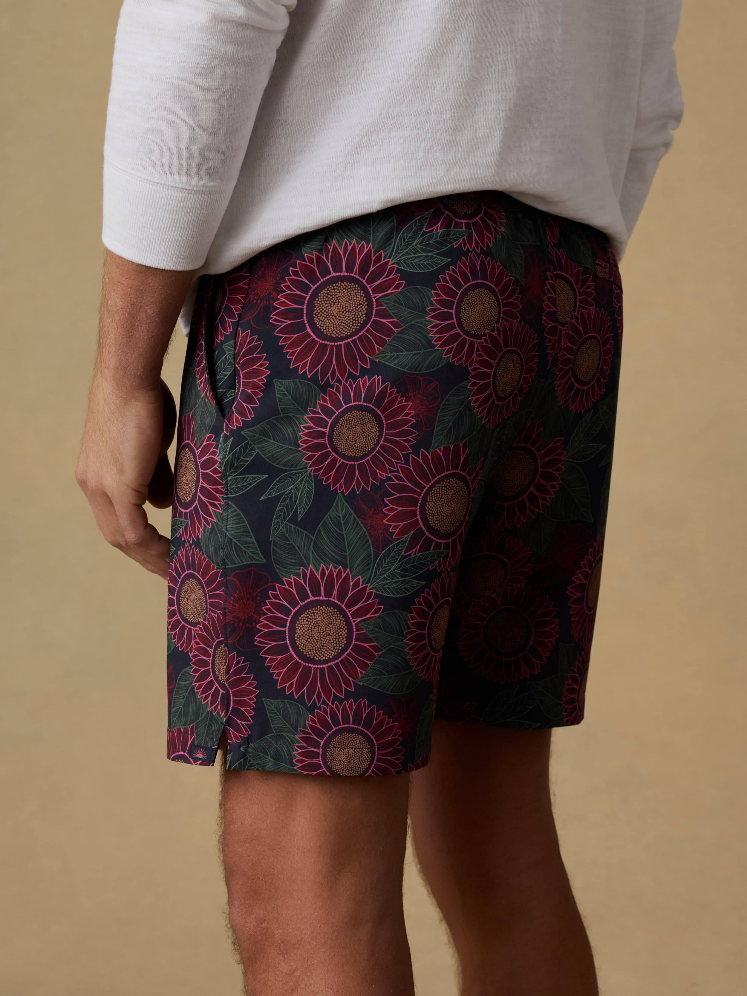 Close up back view of a man wearing a white hoodie and patterned swim trunks against a beige background
