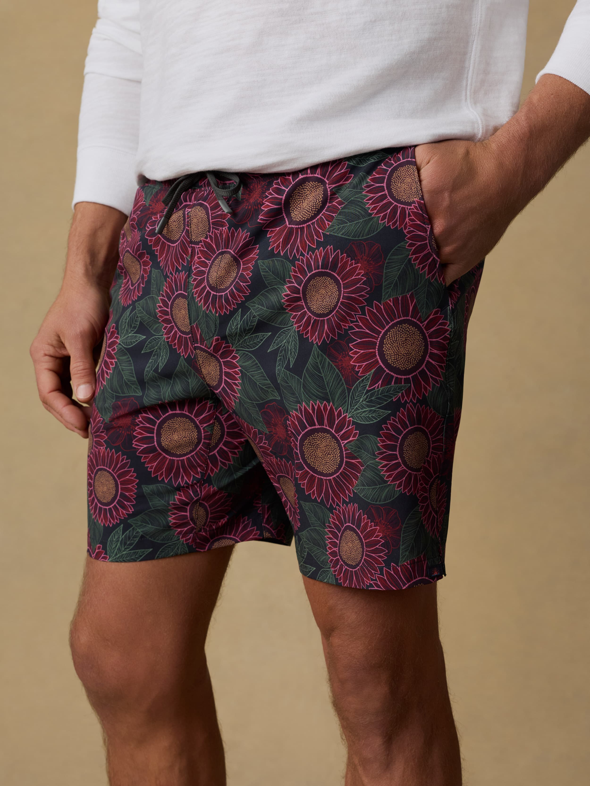 Close up shot of a man wearing a white hoodie and patterned swim trunks against a beige background