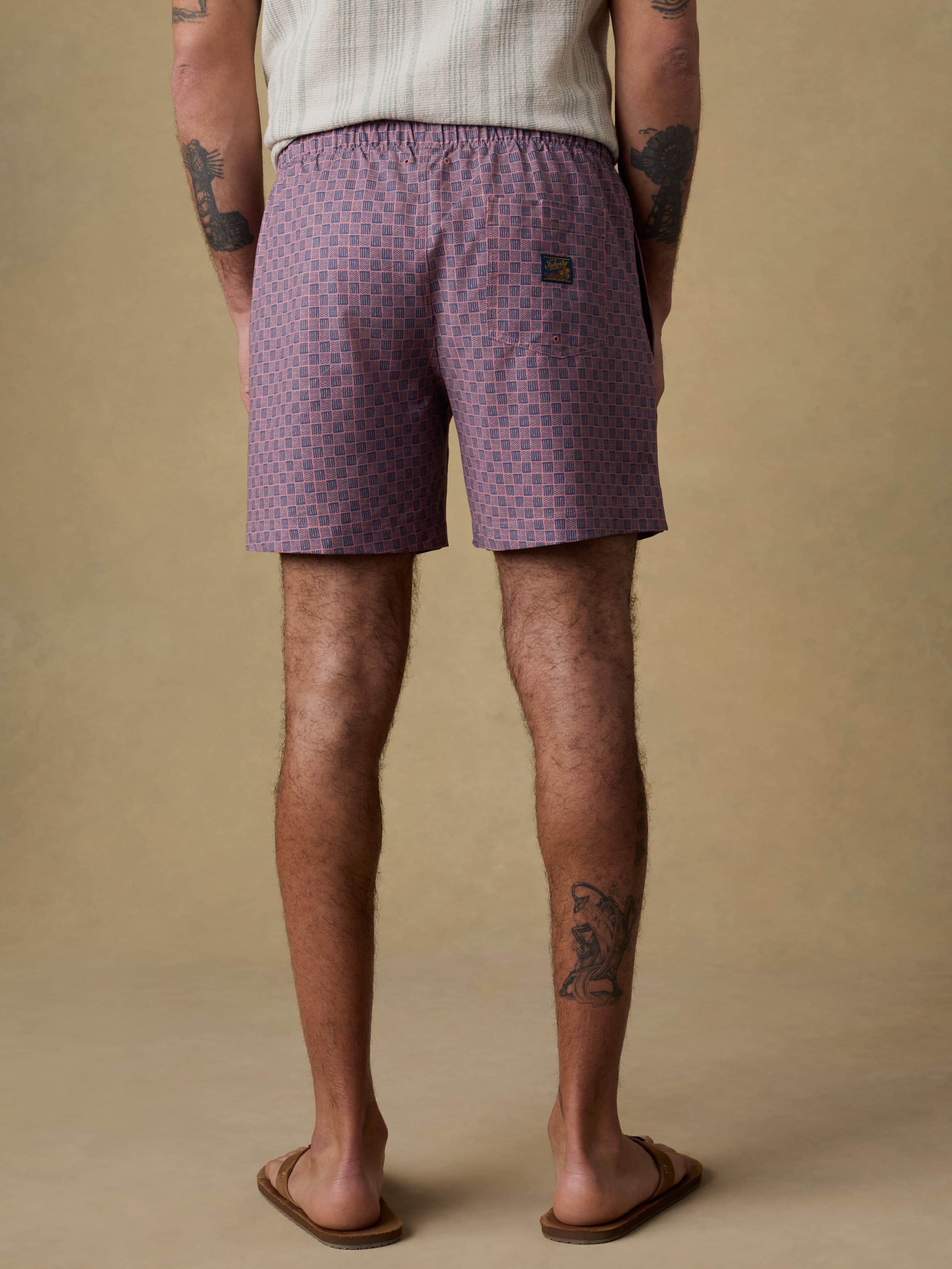 Back view of a man wearing an ivory and green striped short sleeve button up shirt and purple swim trunks against a beige background