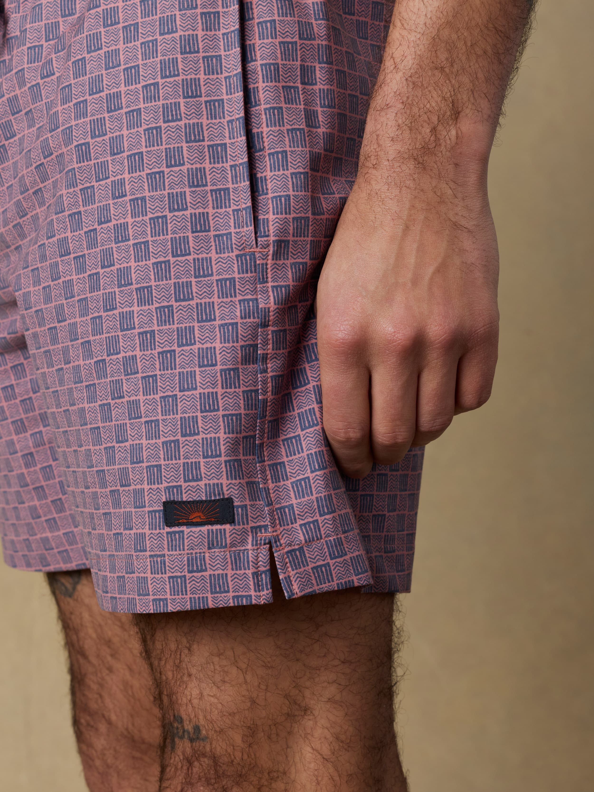 Close-up of a person wearing purple checkered swim trunks with a visible Faherty brand logo near the hem against a beige background
