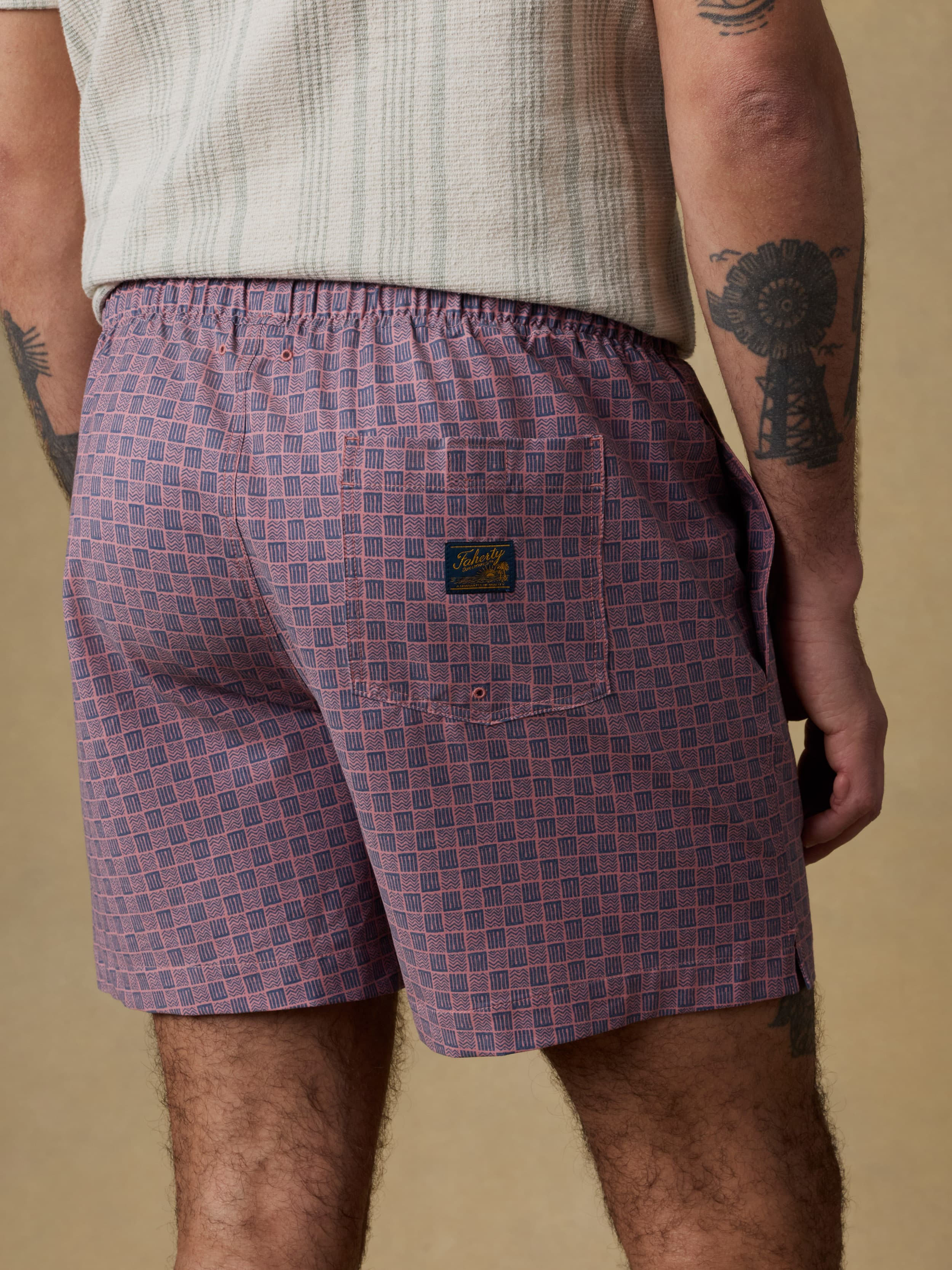 Back view of a man wearing an ivory and green striped short sleeve button up shirt and purple checkered swim trunks against a beige background