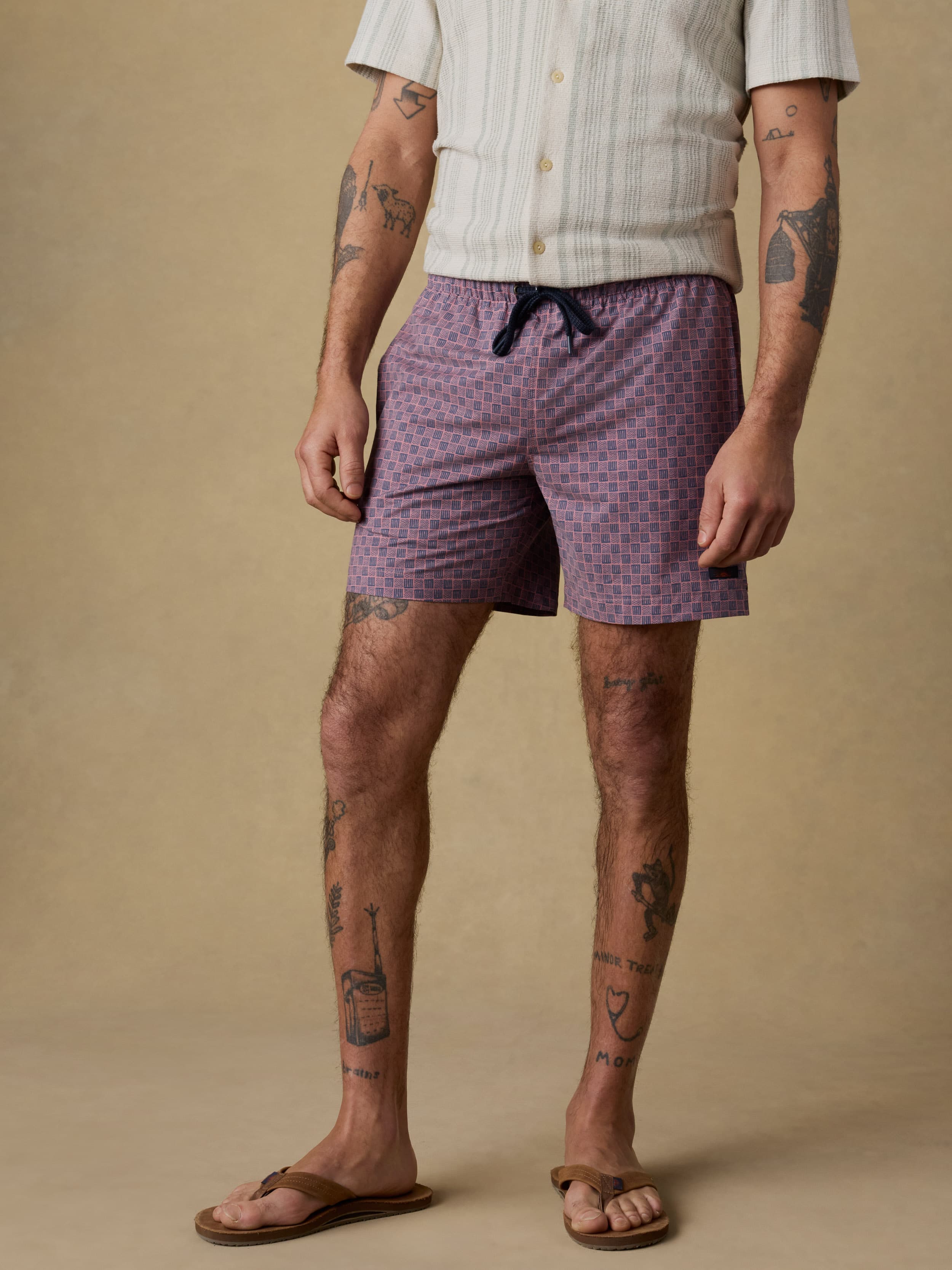 Man wearing an ivory and green striped short sleeve button up shirt and purple swim trunks against a beige background
