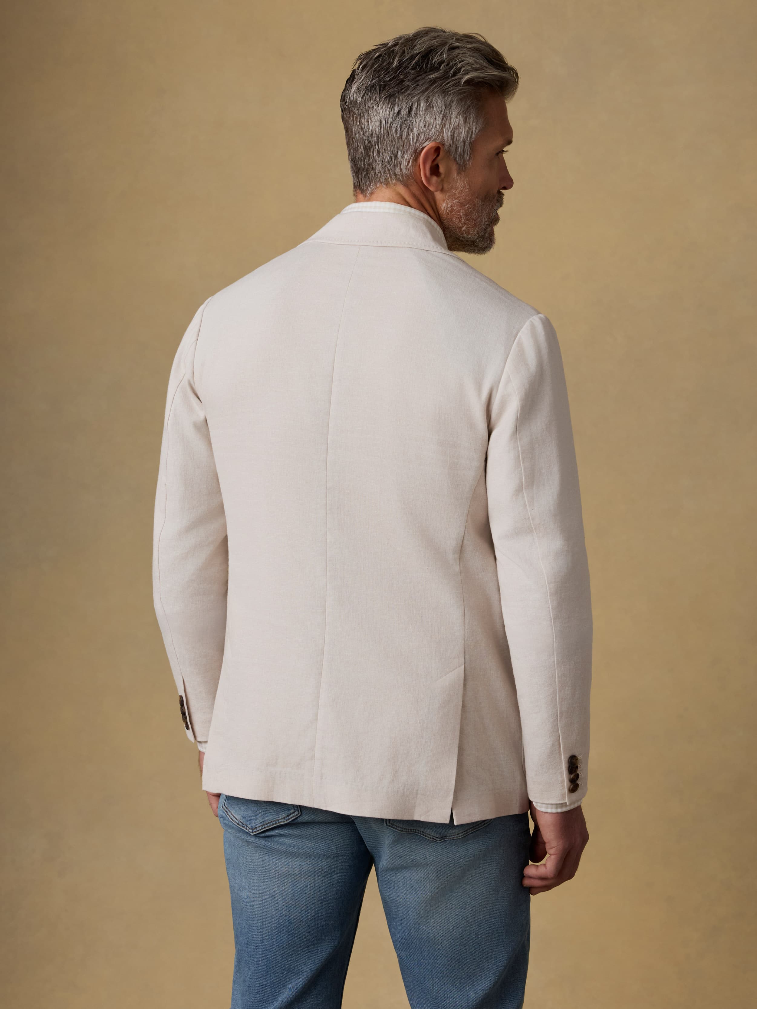Back view of a man wearing a beige blazer and blue jeans against a beige background