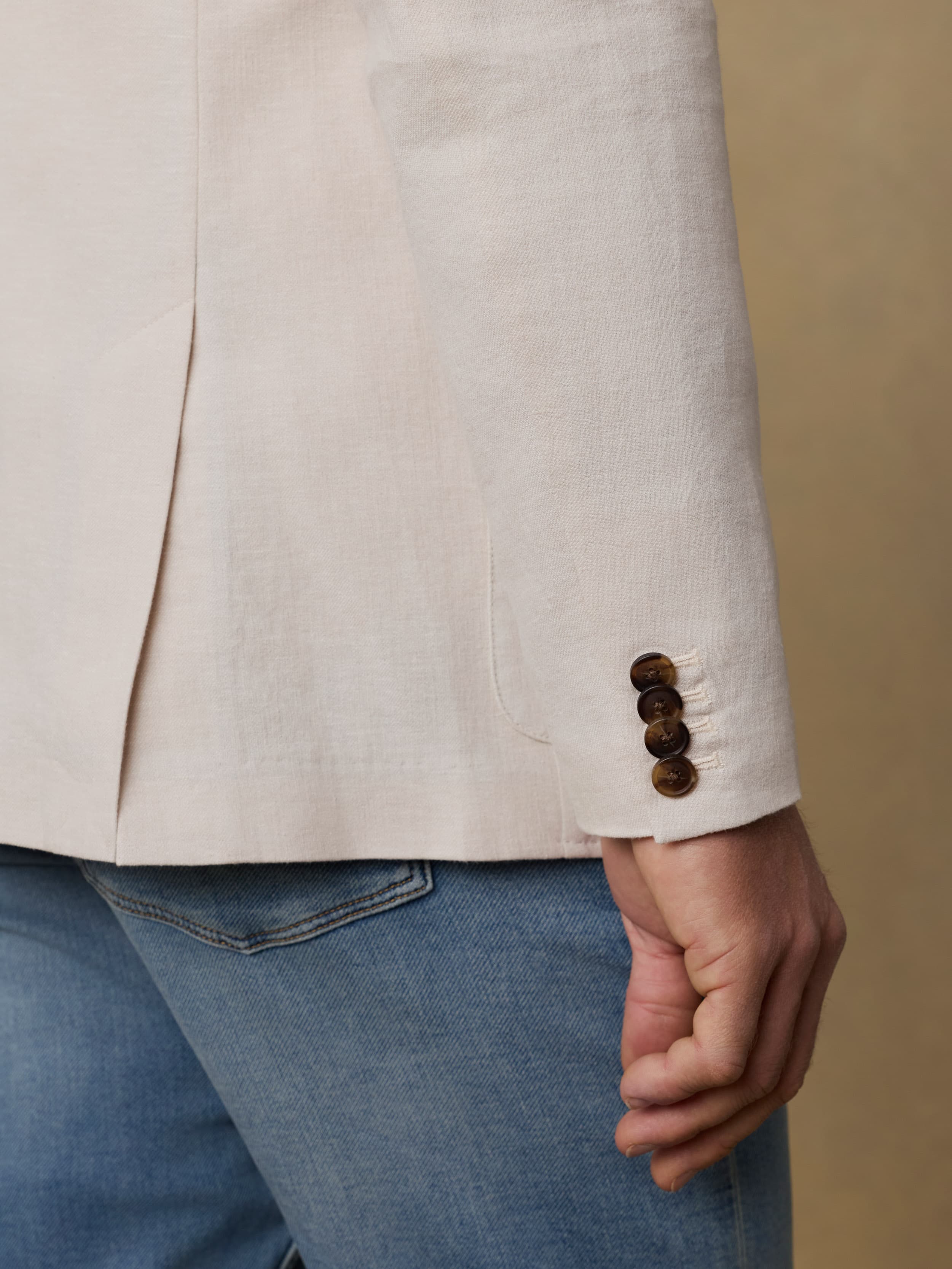 Close-up of a man wearing a beige blazer with brown buttons and blue jeans against a beige background.