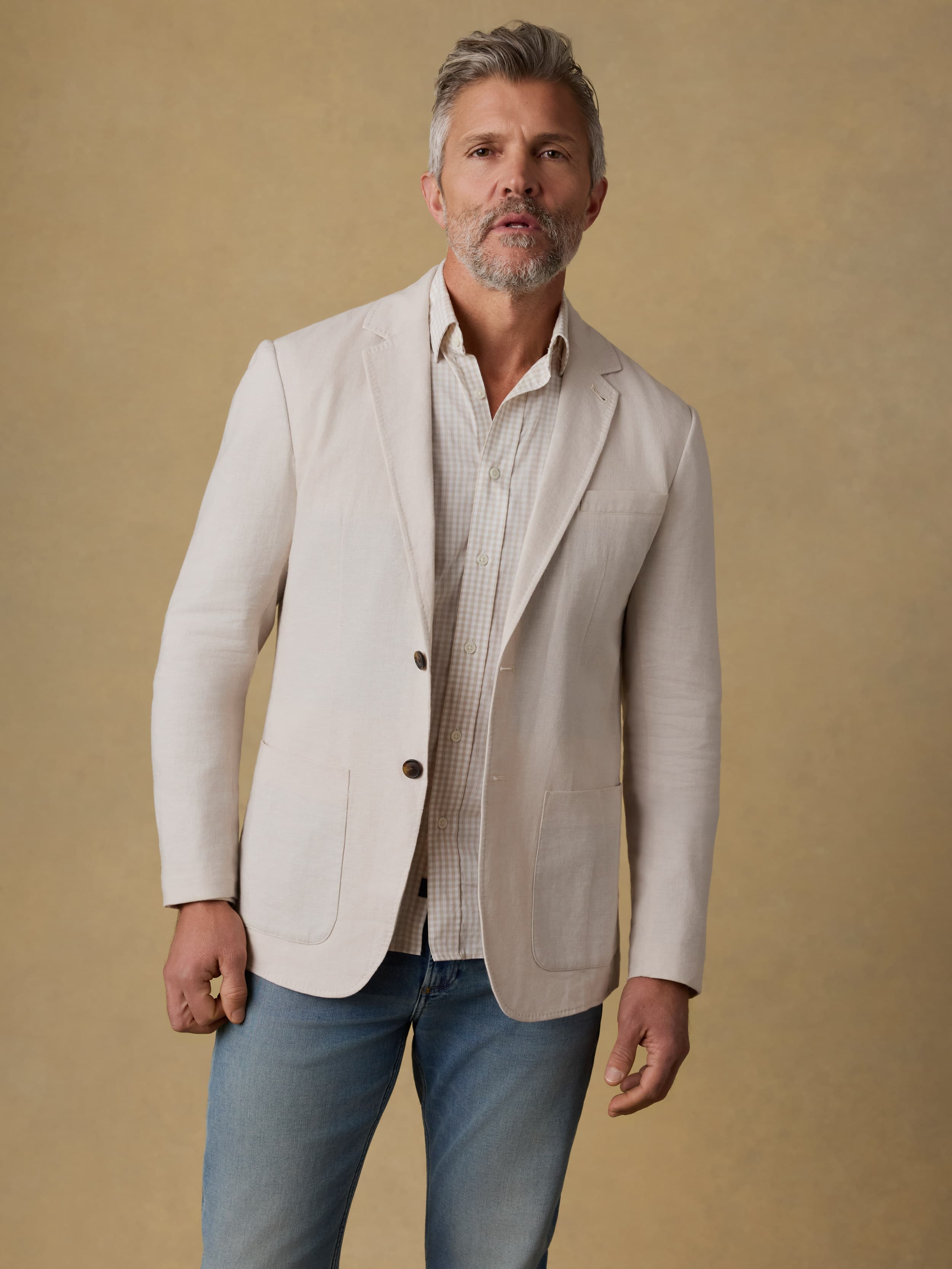 Man wearing a beige blazer over a checkered shirt with blue jeans on a beige background
