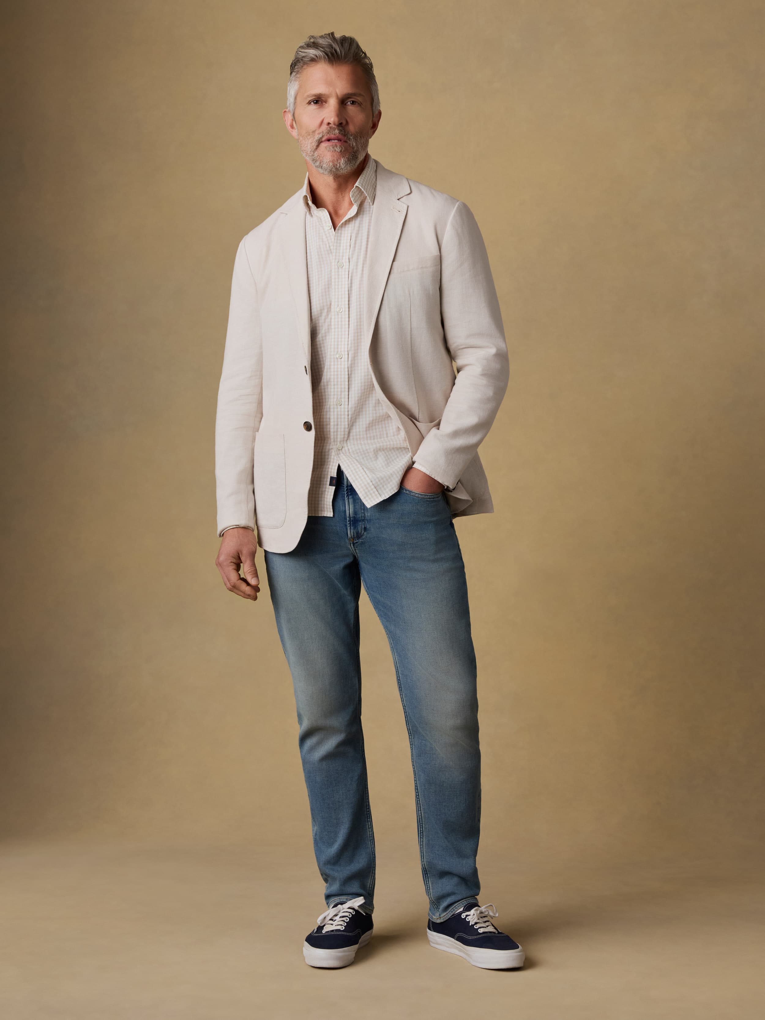 Man wearing a light-colored blazer, checkered shirt, and blue jeans against a beige background