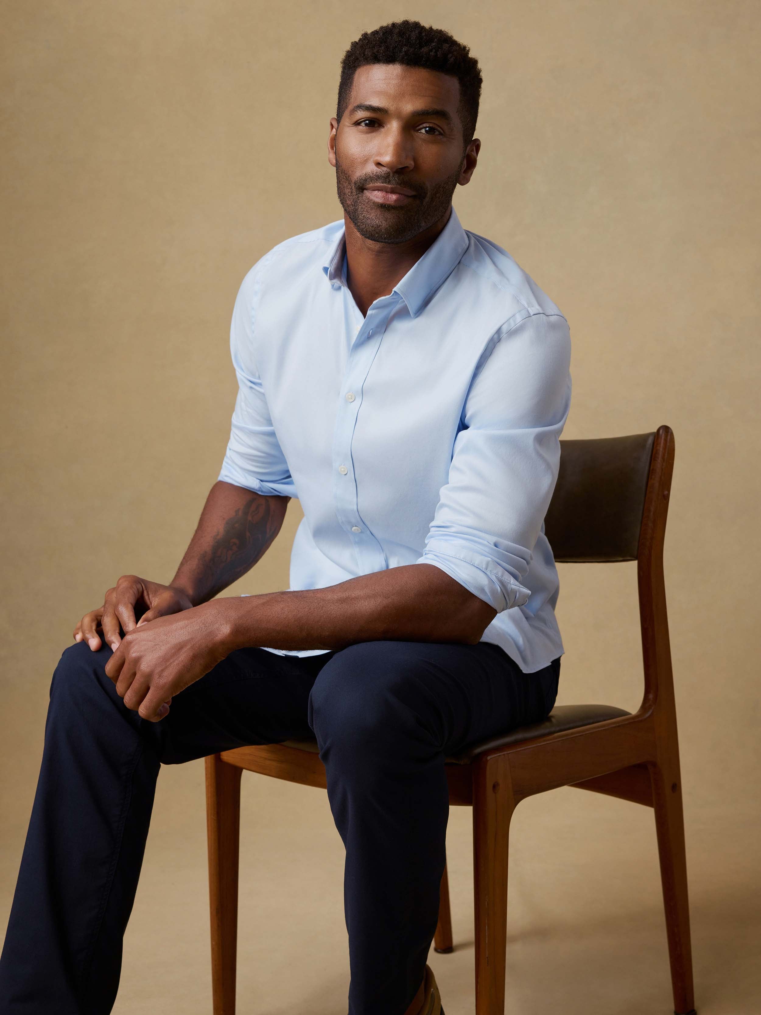 Man sitting in a chair wearing a light blue button-up shirt and navy pants against a beige background