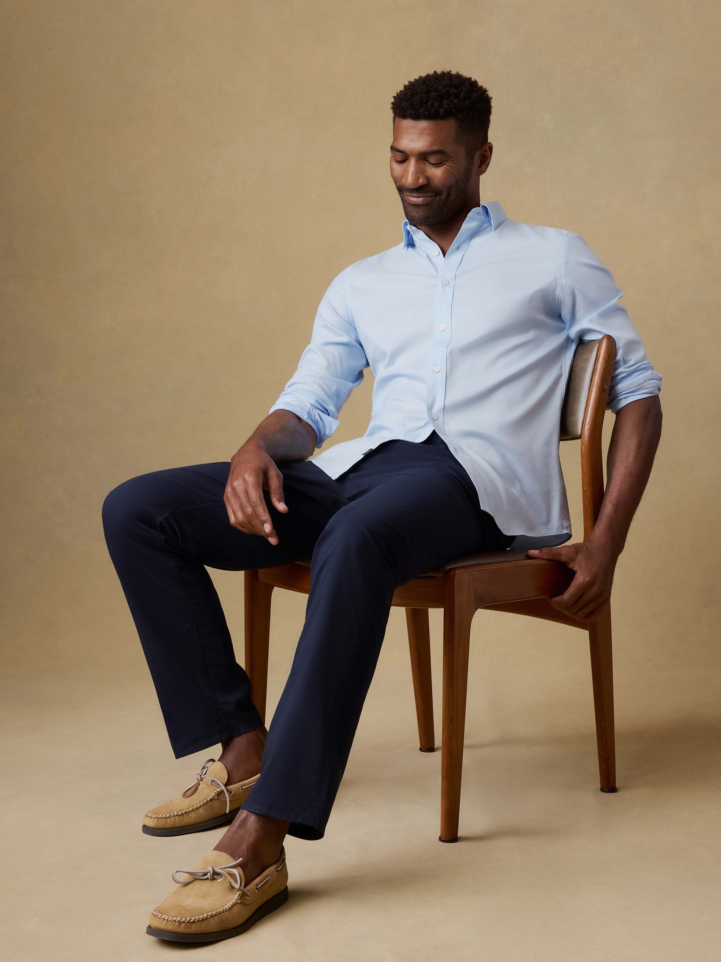Man sitting in a chair wearing a light blue button-up shirt and navy pants against a beige background