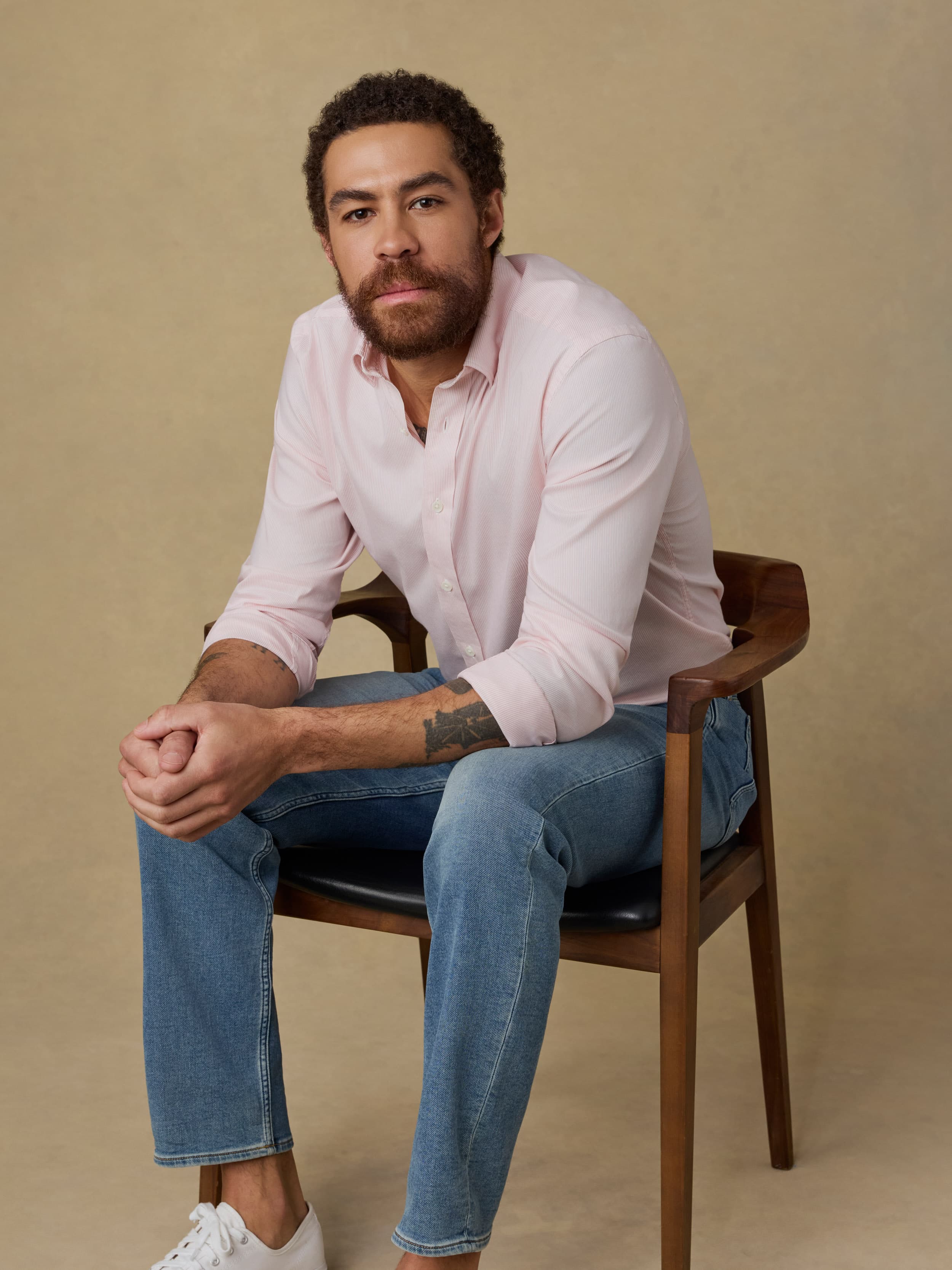 Man sitting on a wooden chair wearing a light pink shirt and blue jeans against a beige background