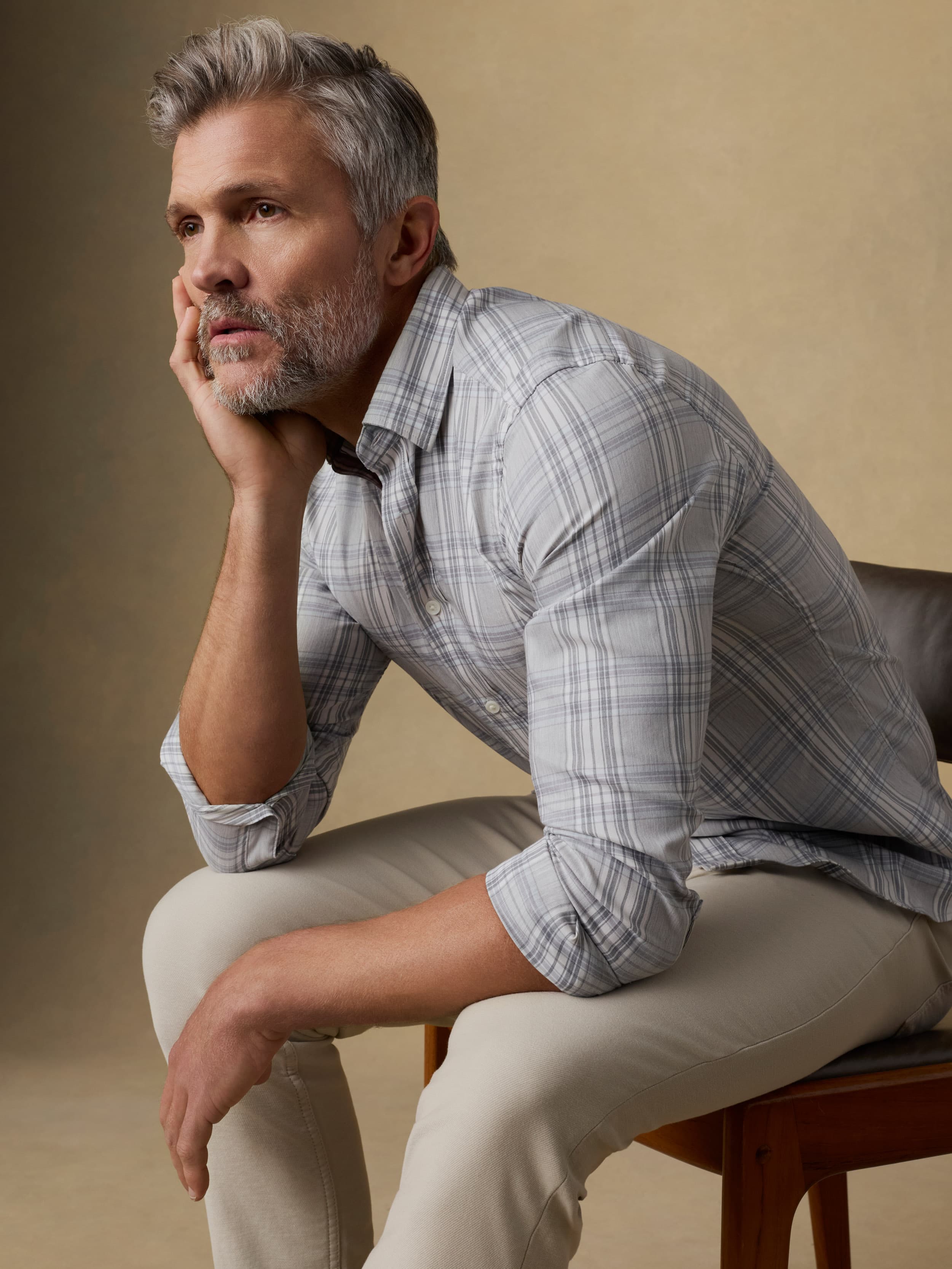 Man sitting on a chair wearing a gray plaid shirt and beige pants against a beige background