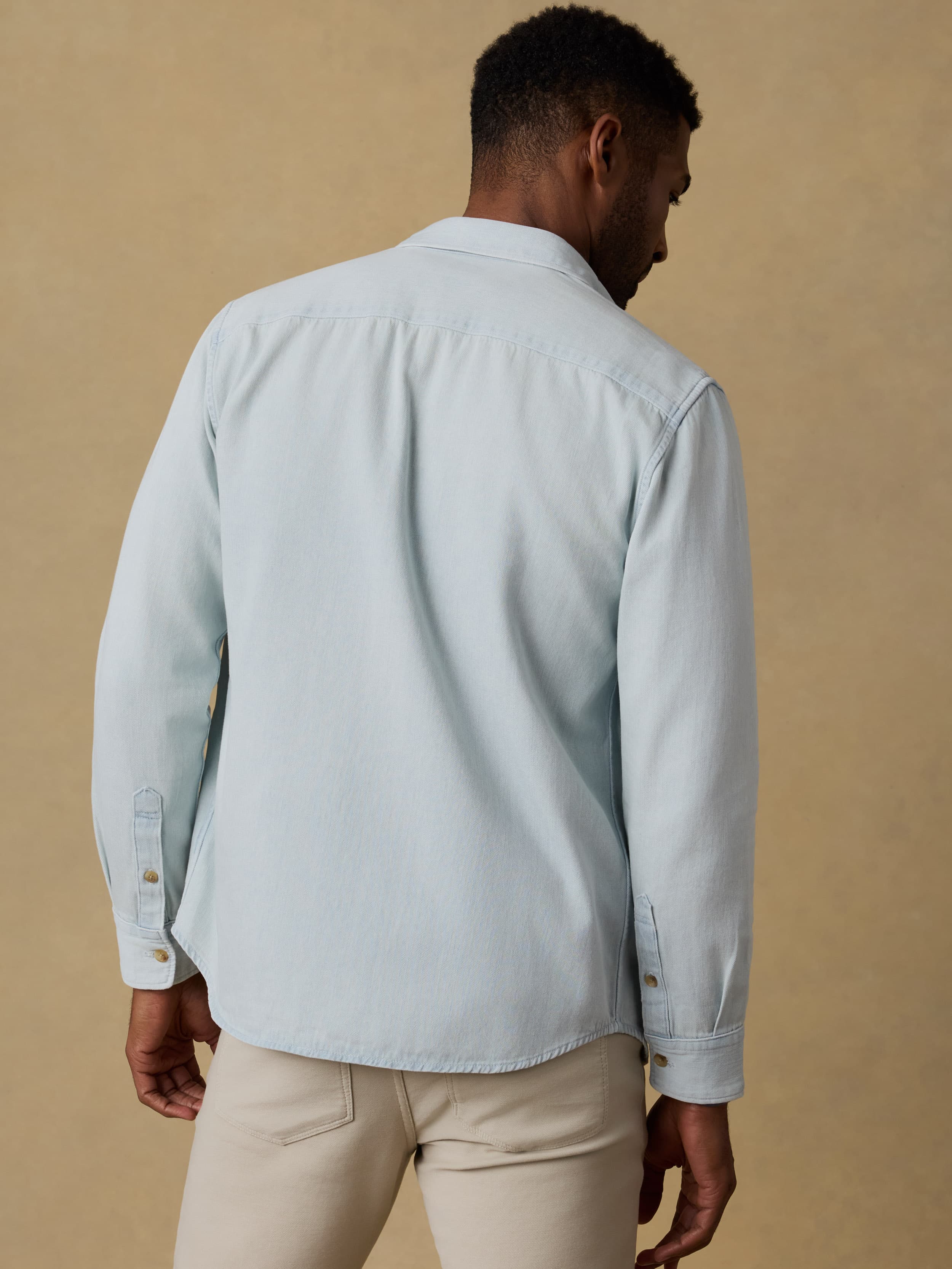 Man wearing a light blue denim shirt and beige jeans against a beige background
