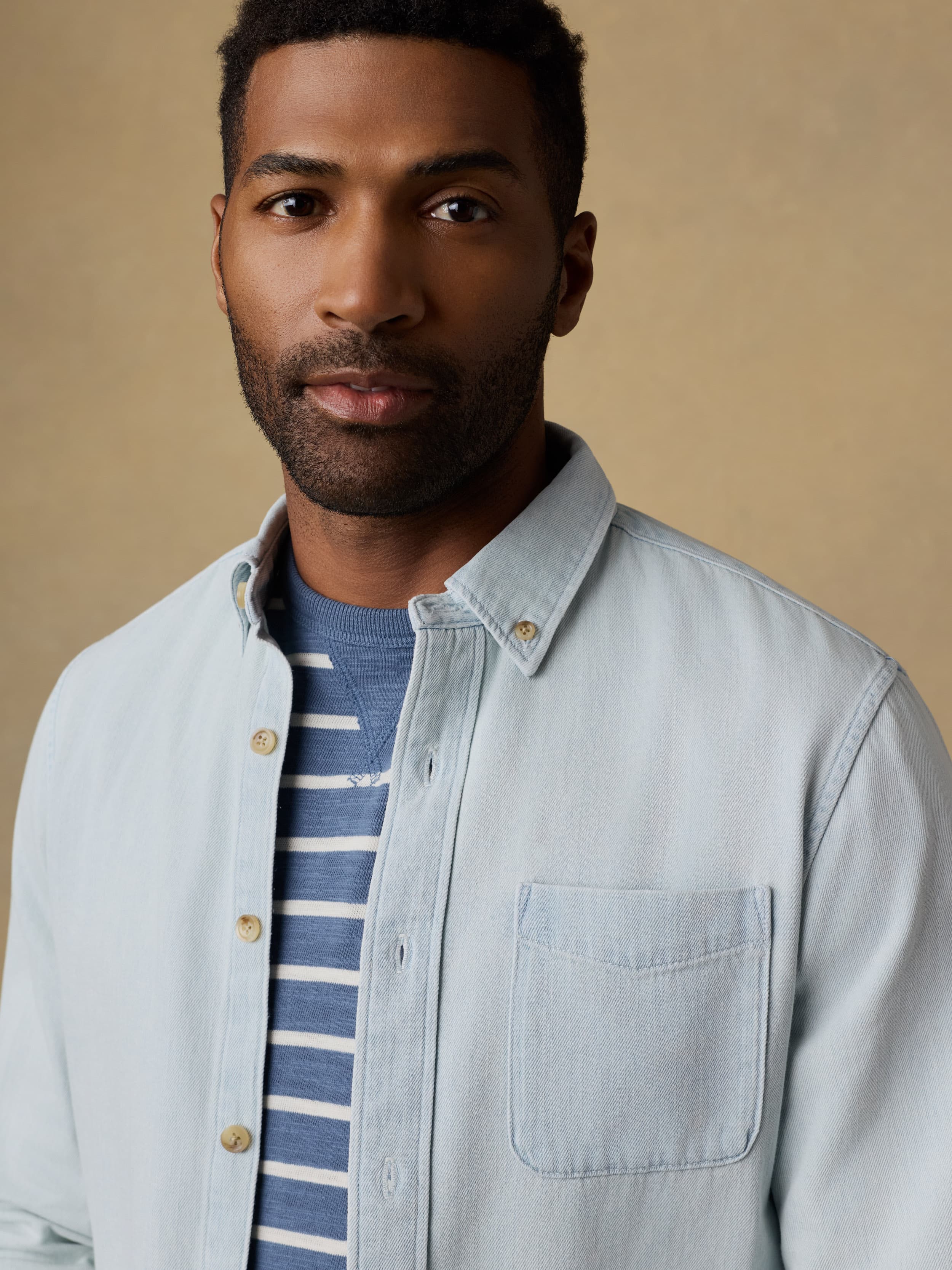 Man wearing a light blue denim shirt over a striped t-shirt against a beige background