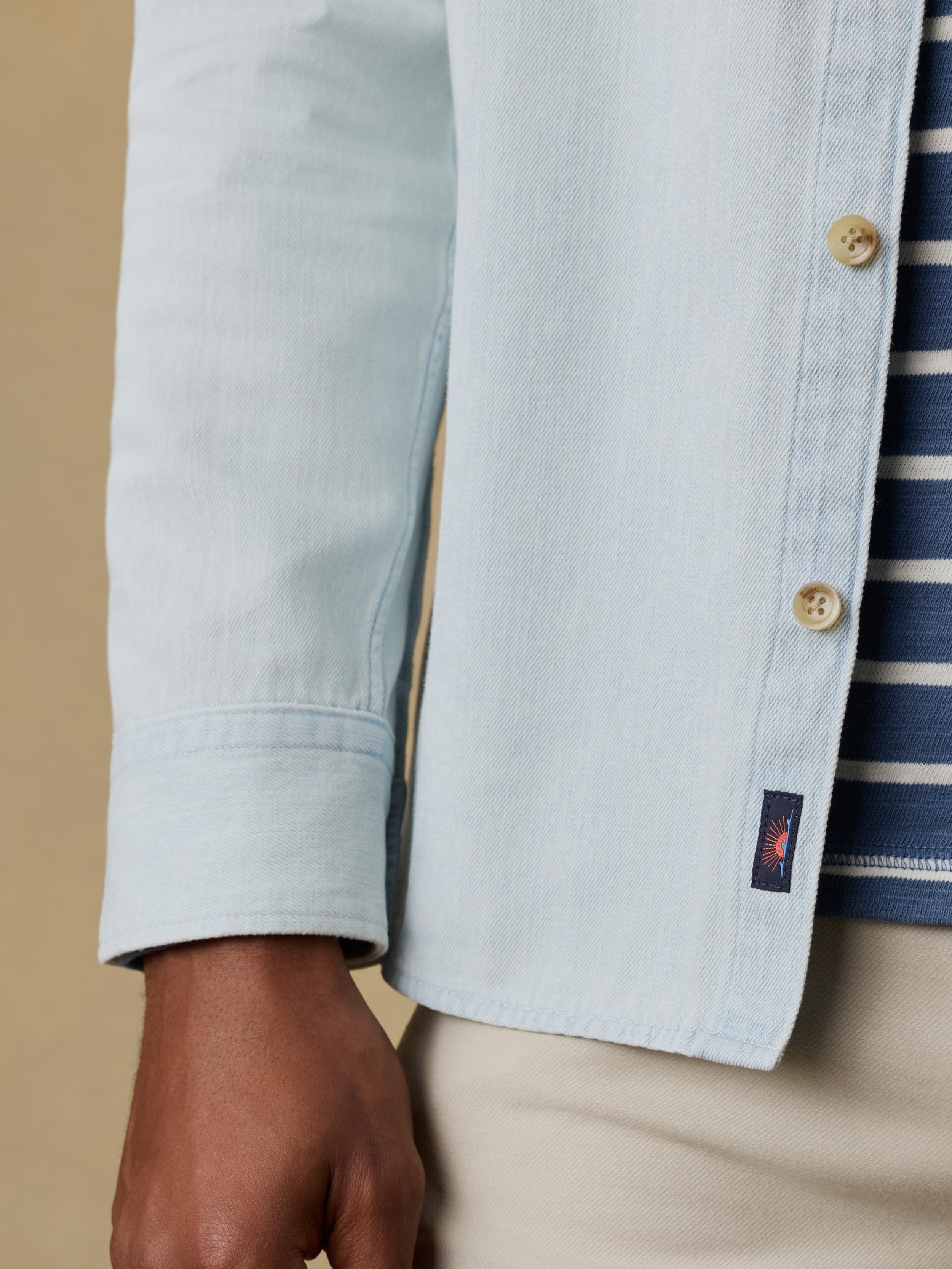 Close up shot of a man wearing a light blue denim shirt over a striped t-shirt and beige jeans against a beige background