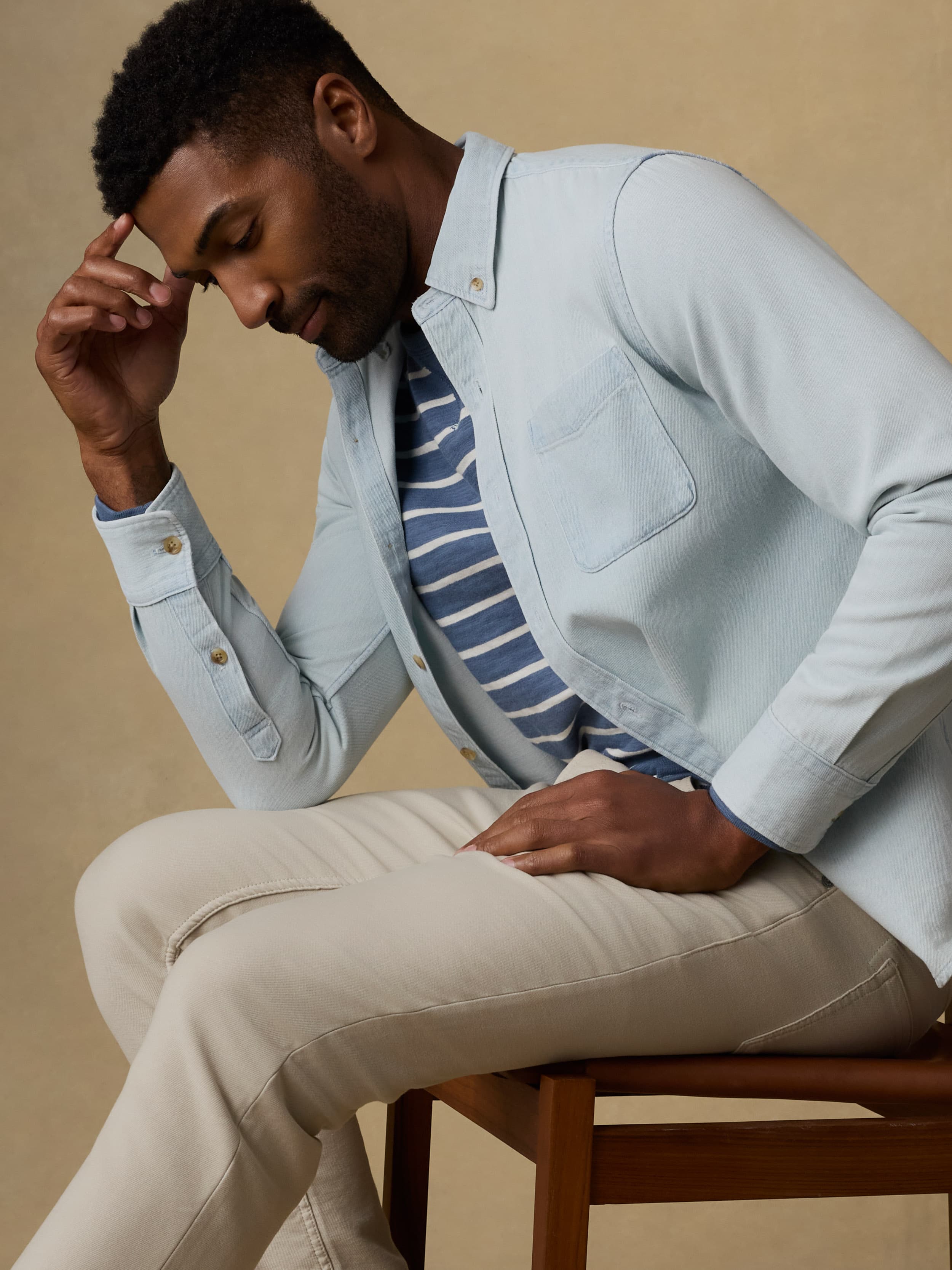 Man wearing a light blue denim shirt over a striped t-shirt and beige jeans against a beige background