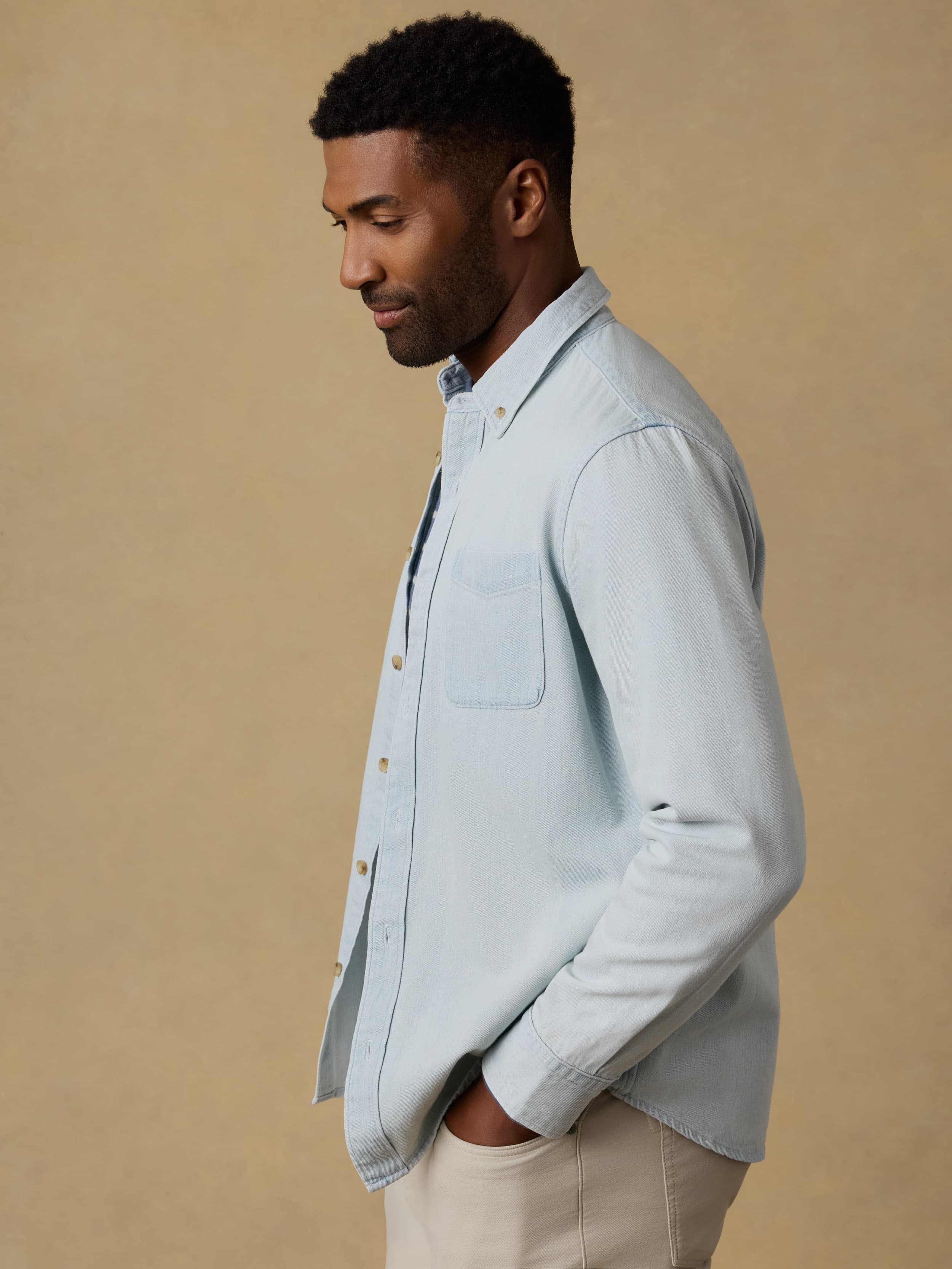 Man wearing a light blue denim shirt over a striped t-shirt and beige jeans against a beige background