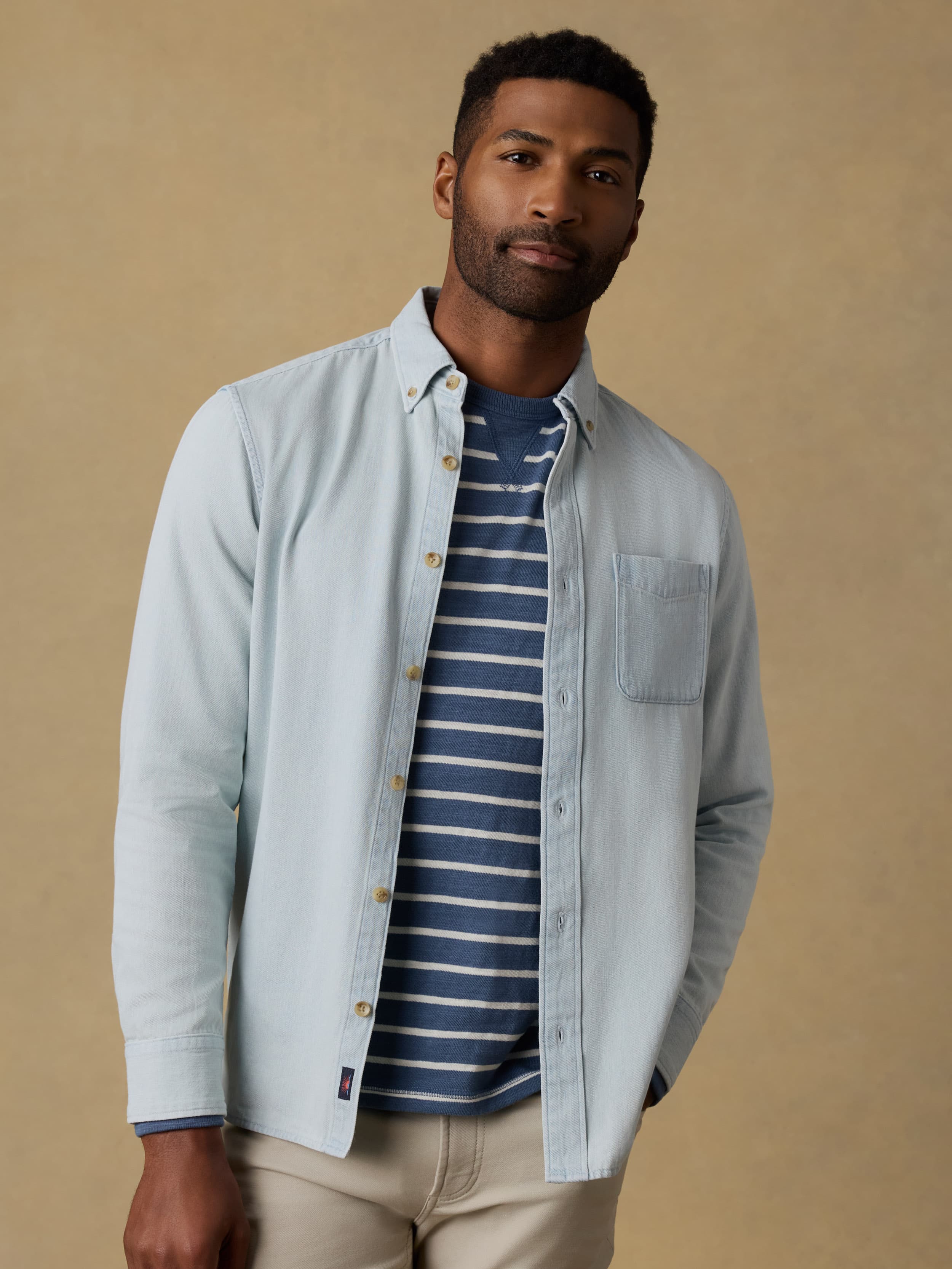 Man wearing a light blue denim shirt over a striped t-shirt and beige jeans against a beige background