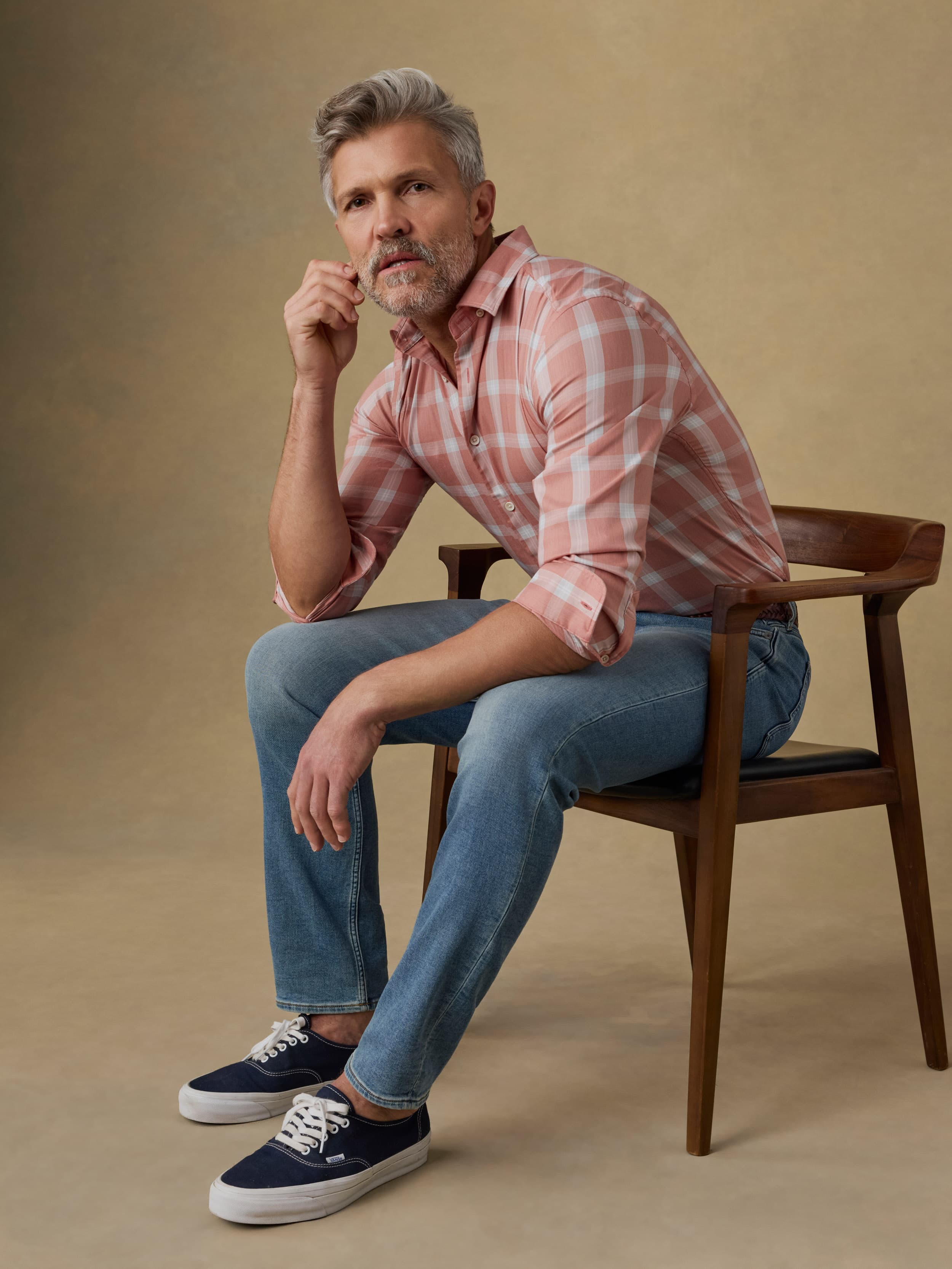 Man sitting on a wooden chair wearing a light red plaid shirt and blue jeans against a beige background