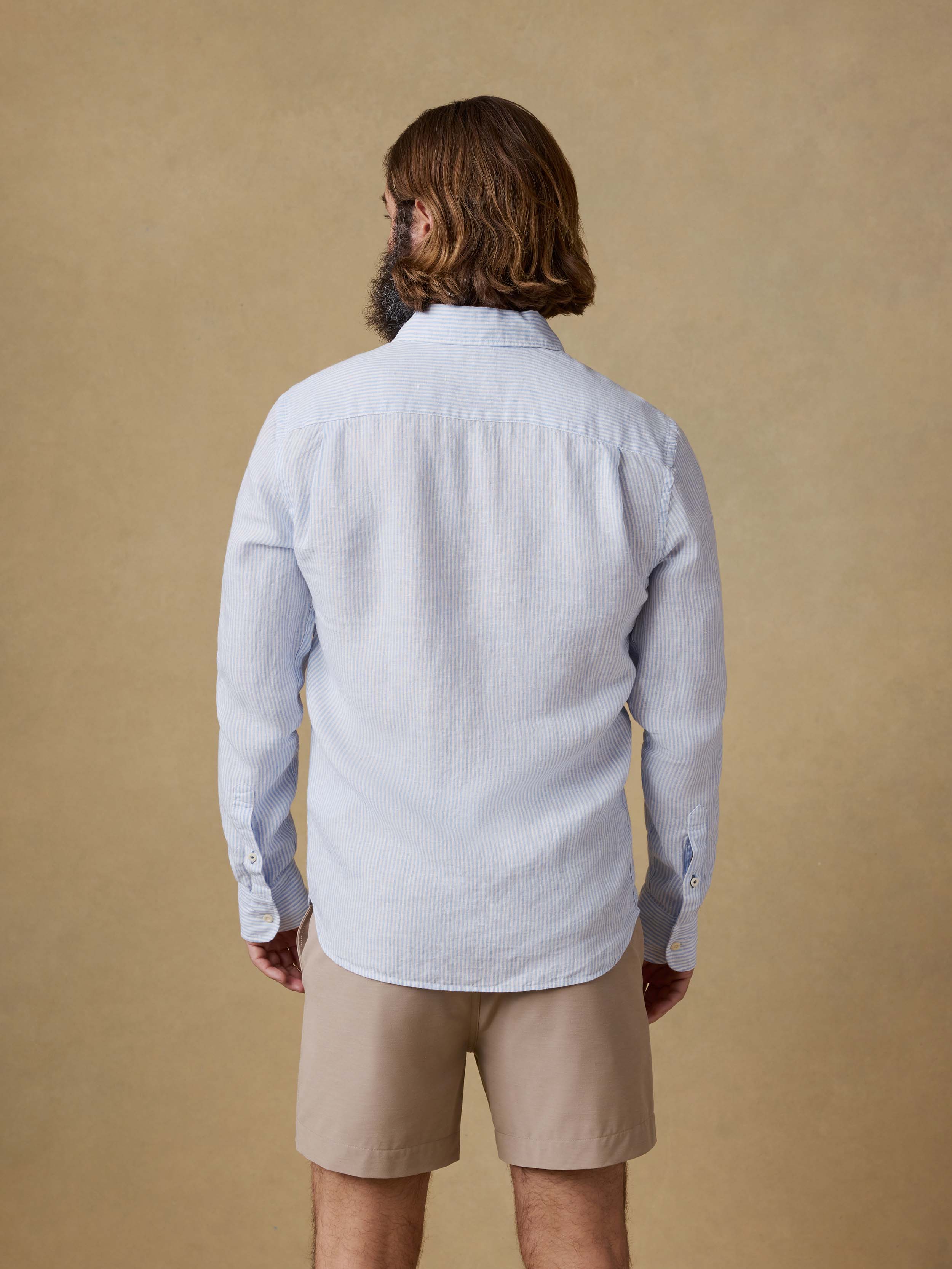Back view of a man wearing a light blue striped button up shirt and beige shorts against a beige background