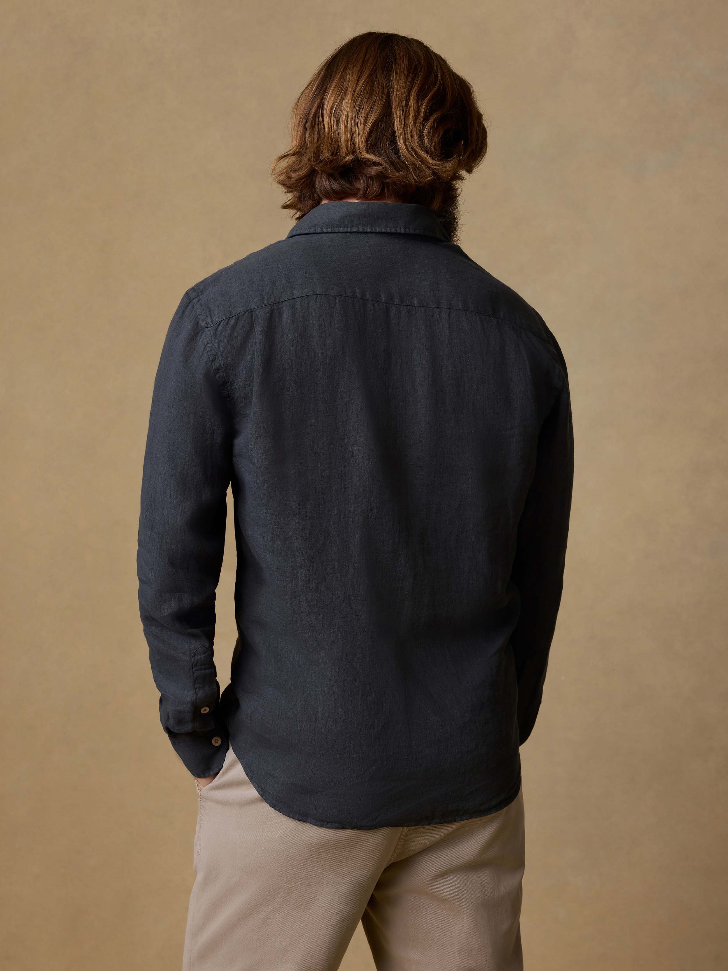 Back view of a man wearing a black button up shirt and beige pants against a beige background