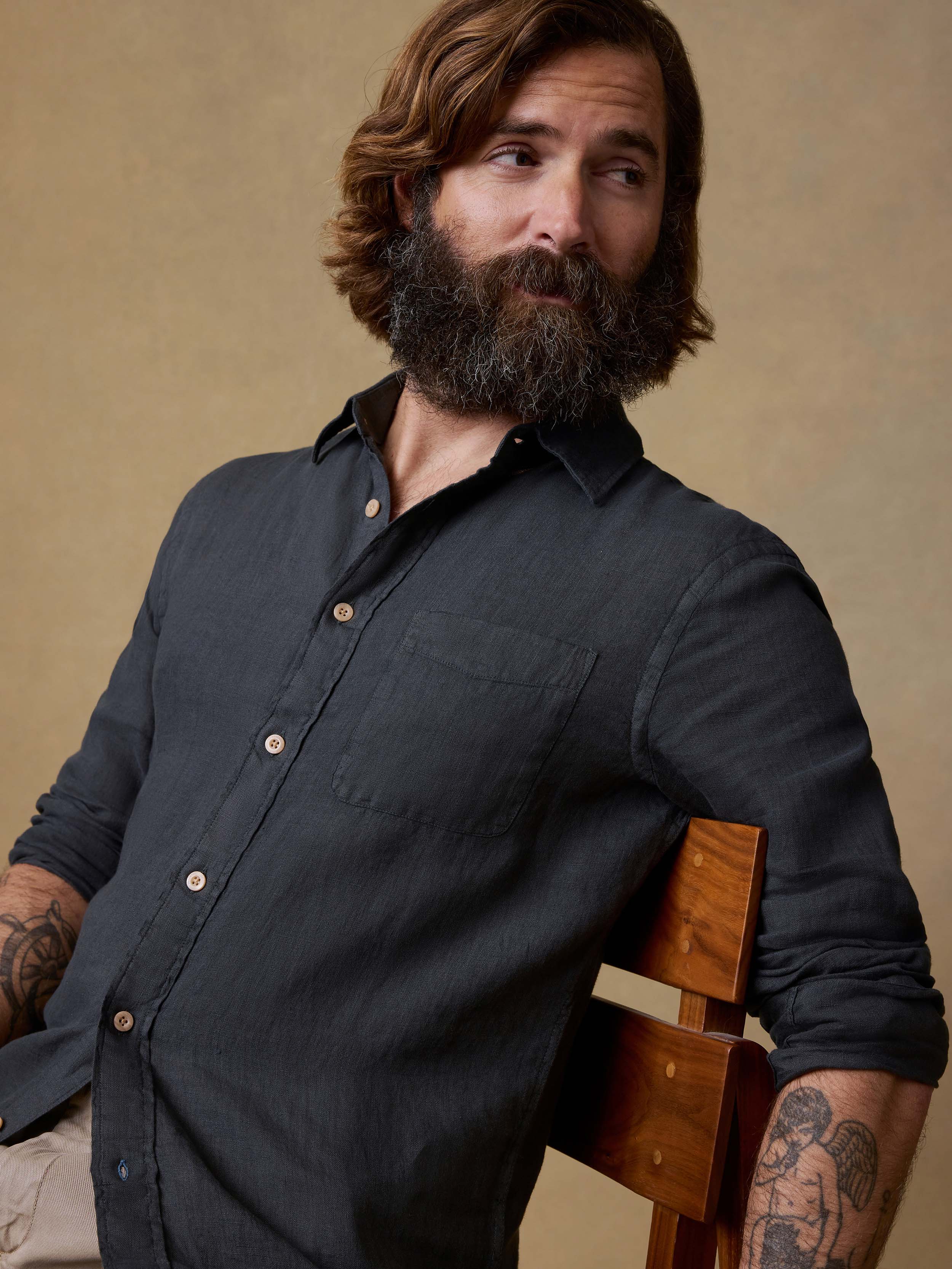 Man sitting in a chair wearing a black button up shirt and beige pants against a beige background