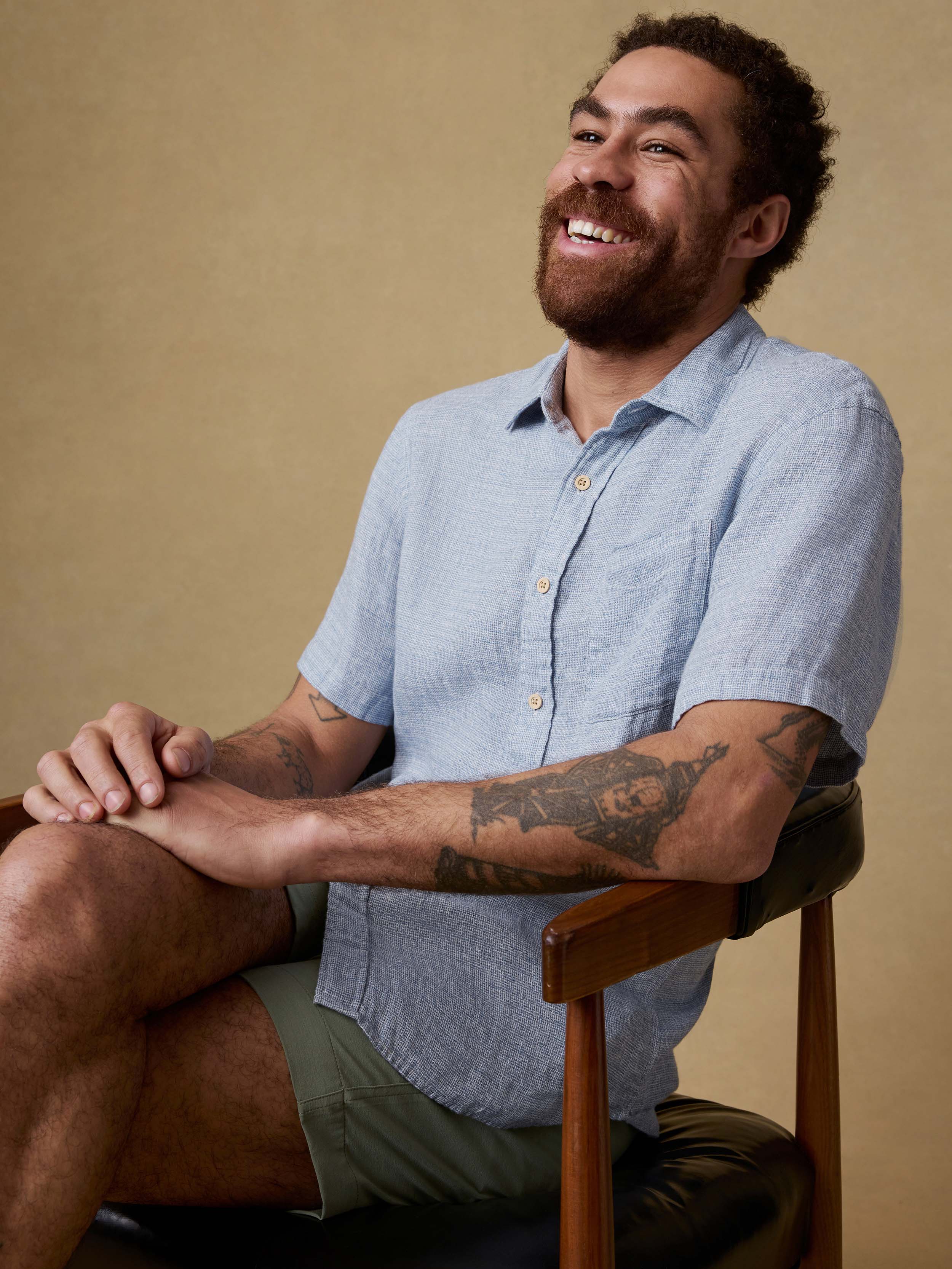 Man sitting on a chair wearing a light blue linen button up shirt and green shorts against a beige background
