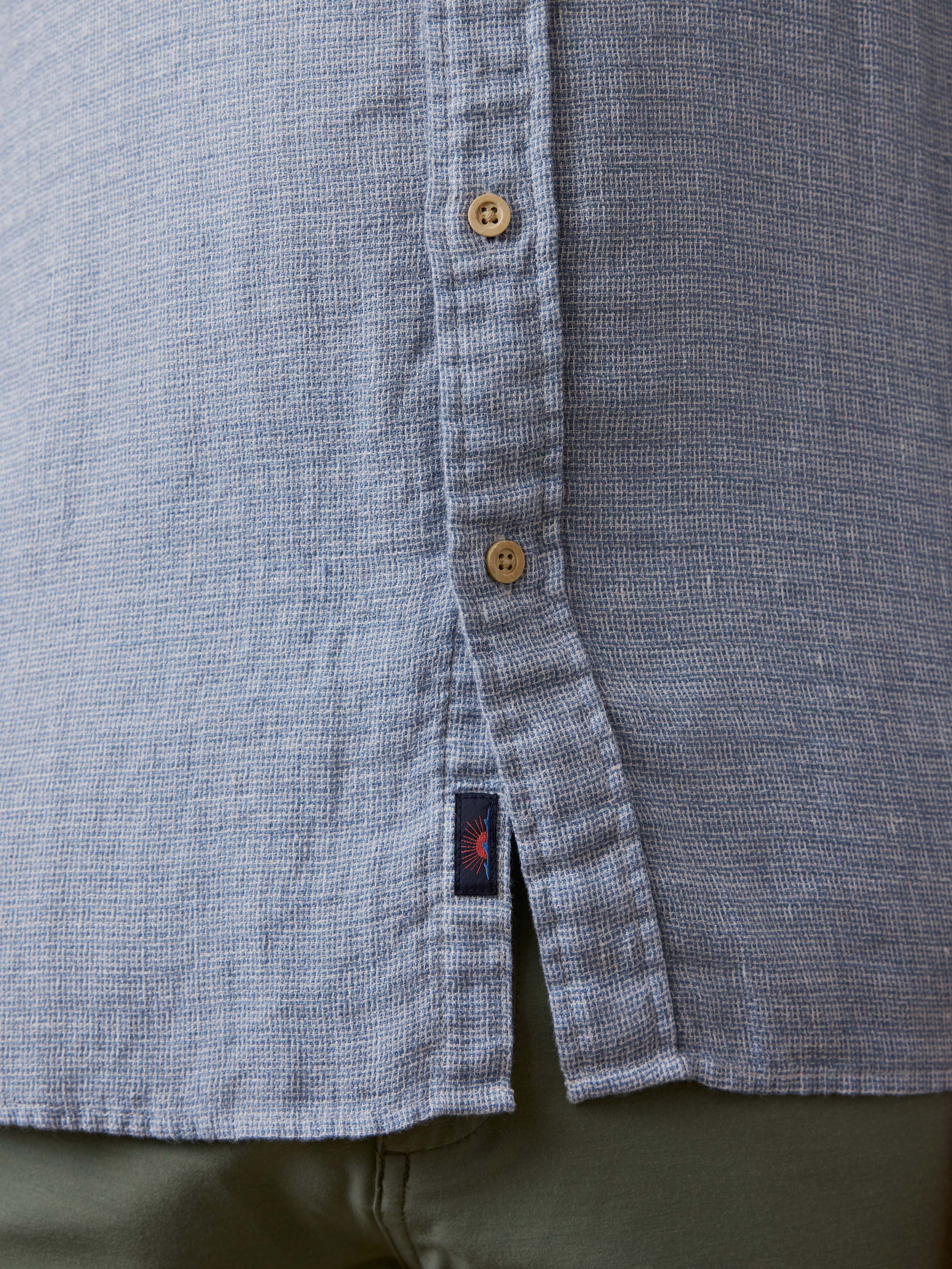 Detail shot of a man wearing a light blue linen button up shirt with visible Faherty brand logo and green shorts 