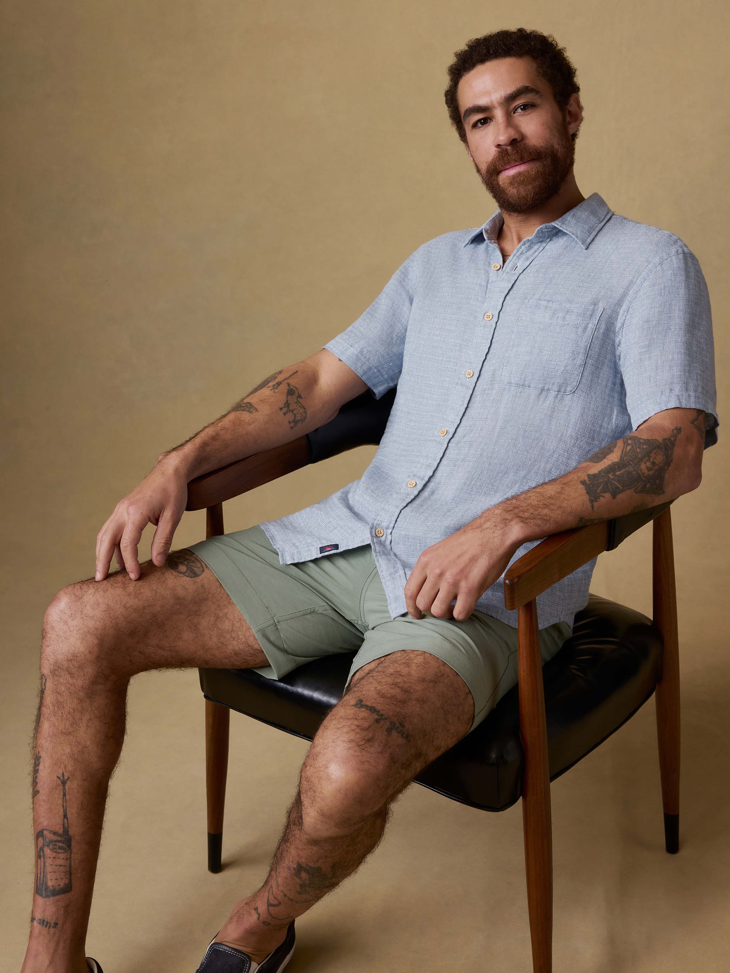 Man sitting on a chair wearing a light blue linen button up shirt and green shorts against a beige background