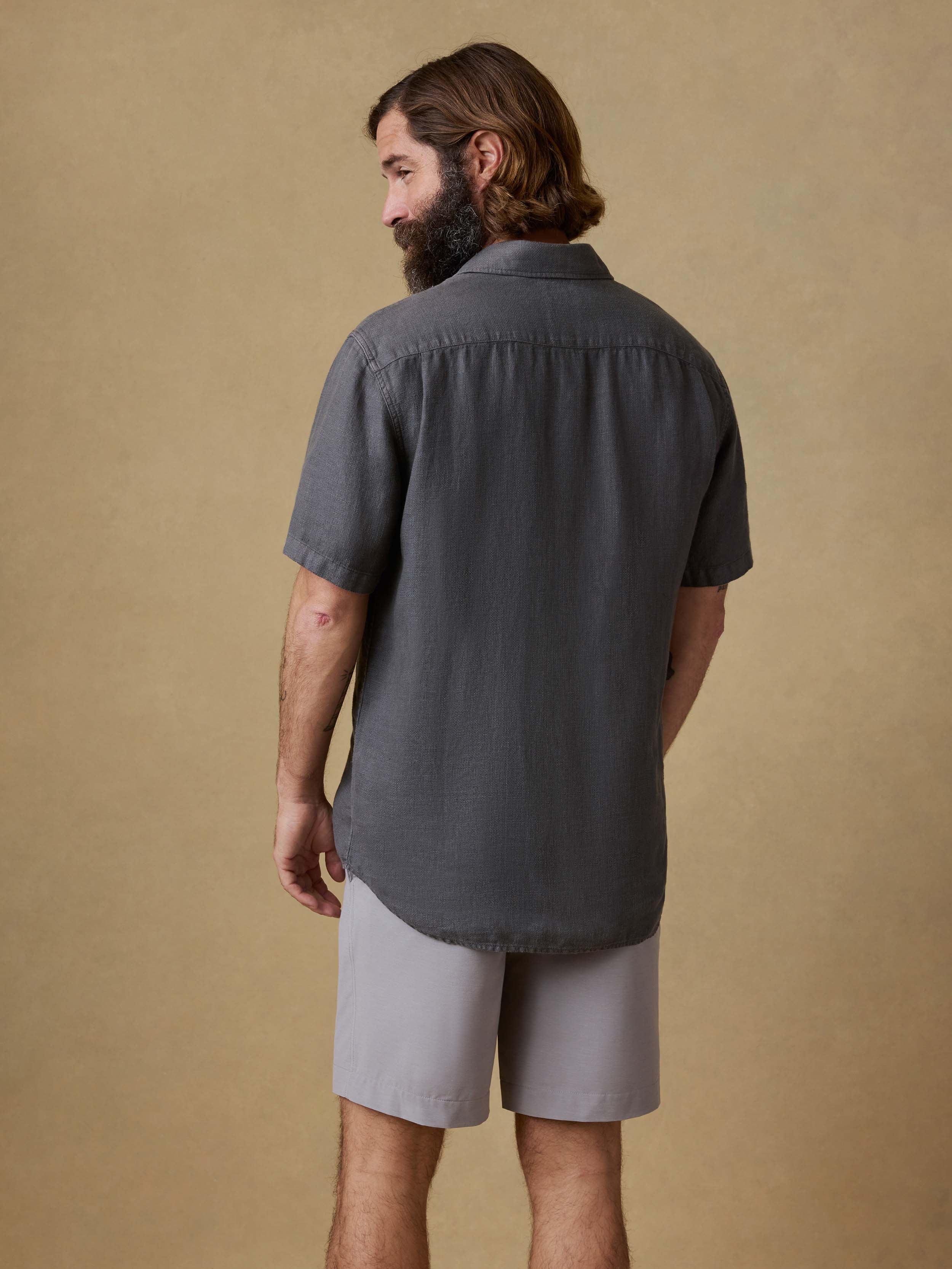 Back view of a man wearing a black short-sleeve linen button up shirt and light gray shorts against a beige background