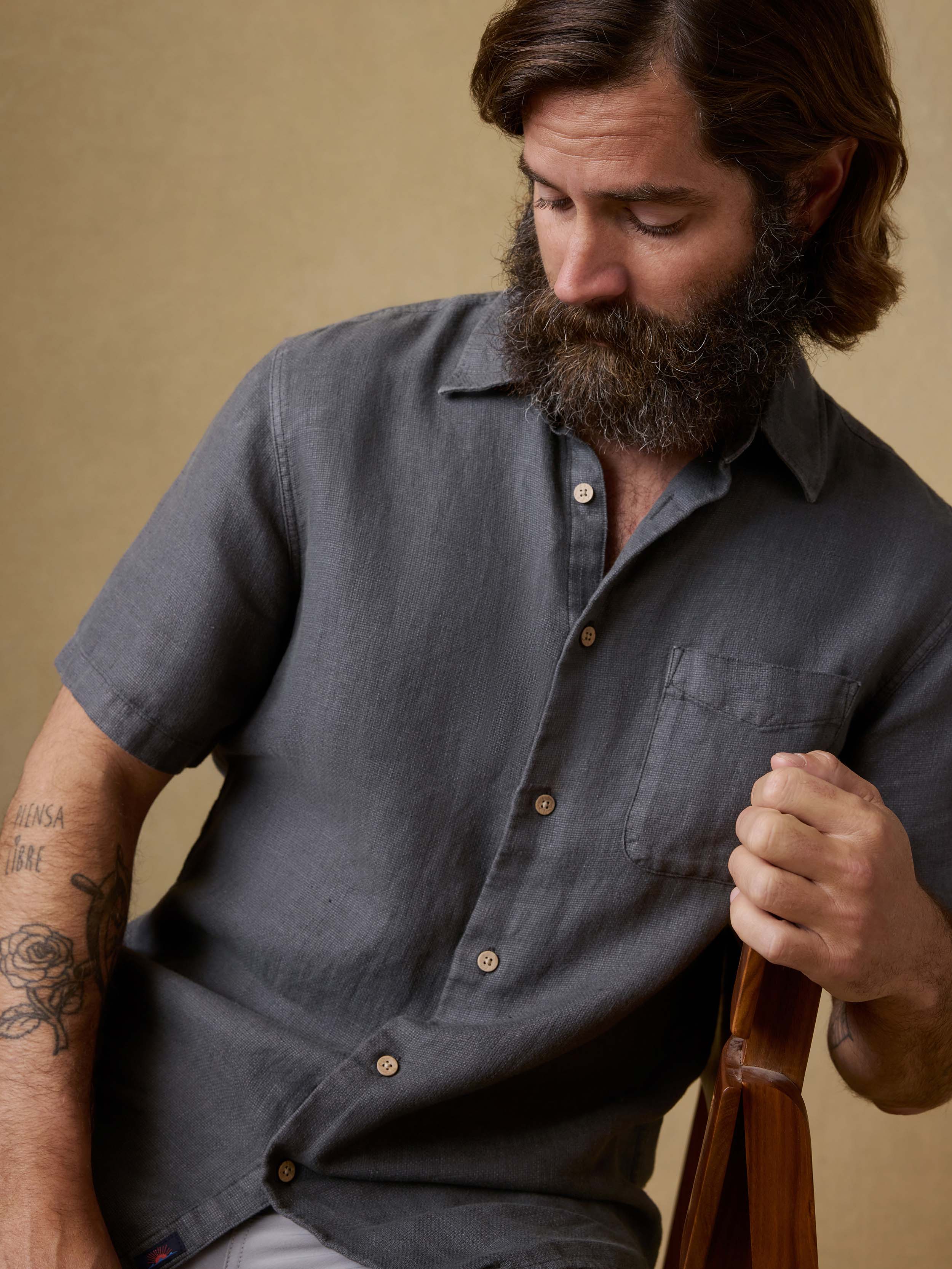 Man sitting in a chair wearing a black short-sleeve linen button up shirt and light gray shorts against a beige background