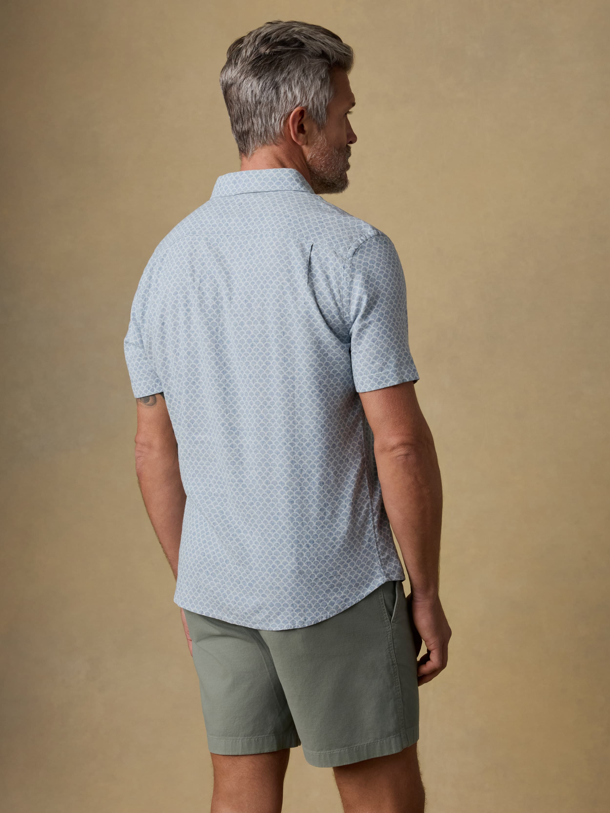 Back view of a man wearing a light blue patterned shirt and green shorts against a beige background