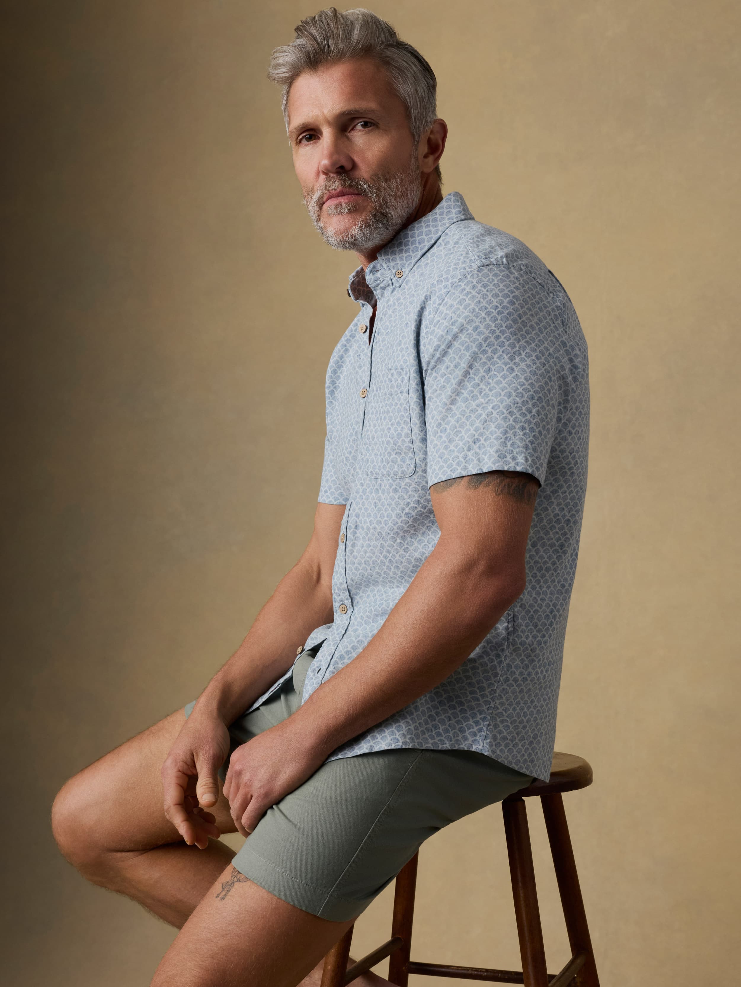 Man sitting on a wooden stool wearing a light blue patterned shirt and green shorts against a beige background