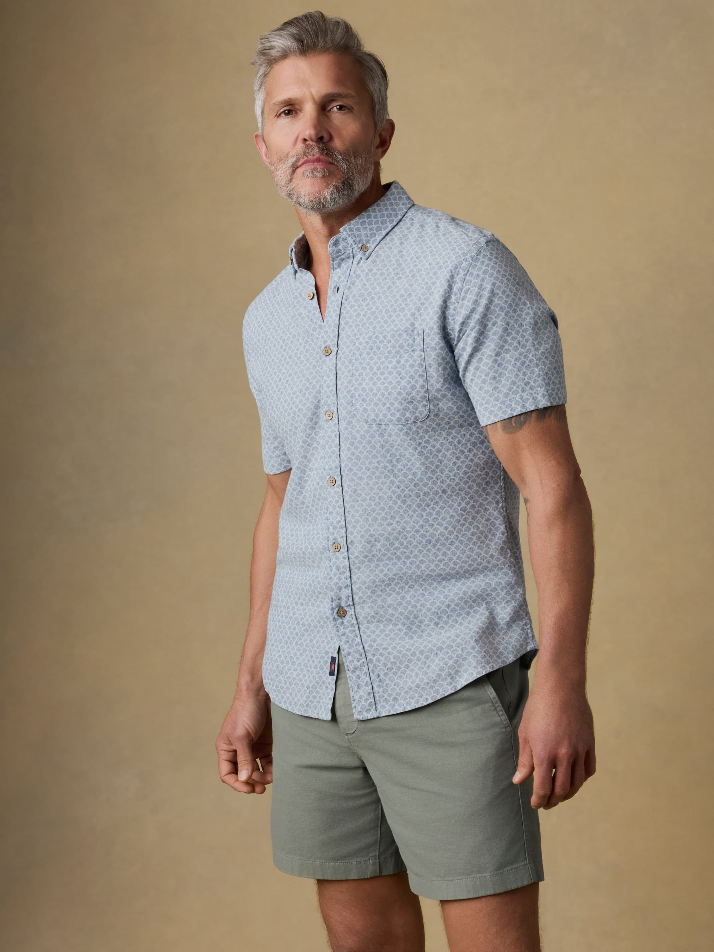 Man wearing a light blue patterned shirt and green shorts against a beige background