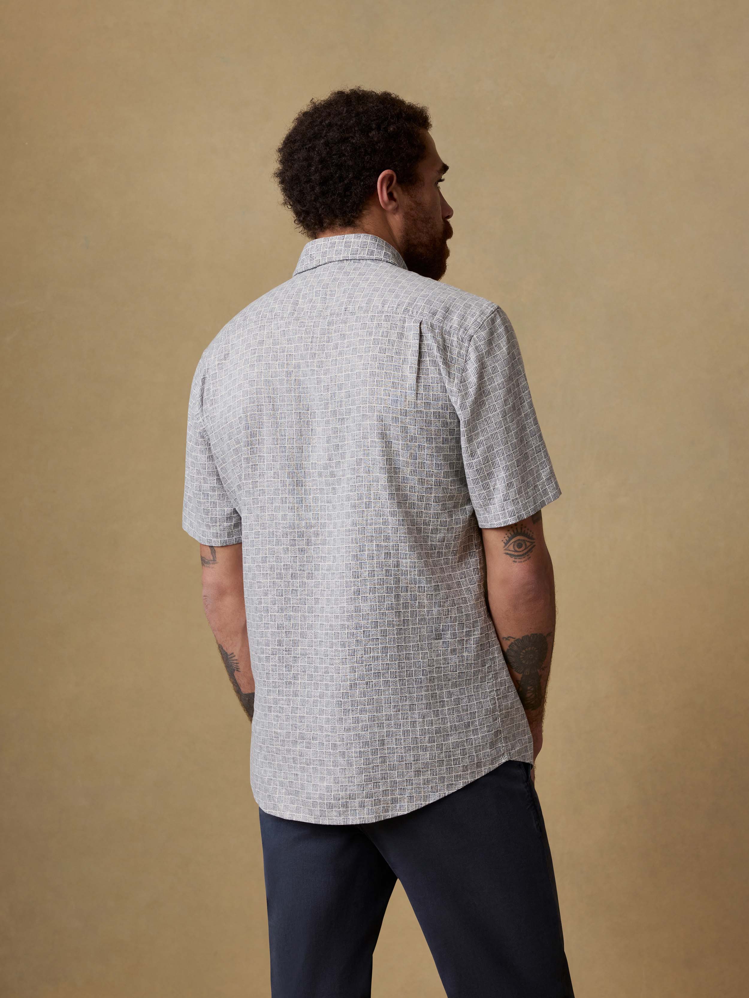 Back view of a man wearing a patterned short-sleeve shirt and navy pants against a beige background