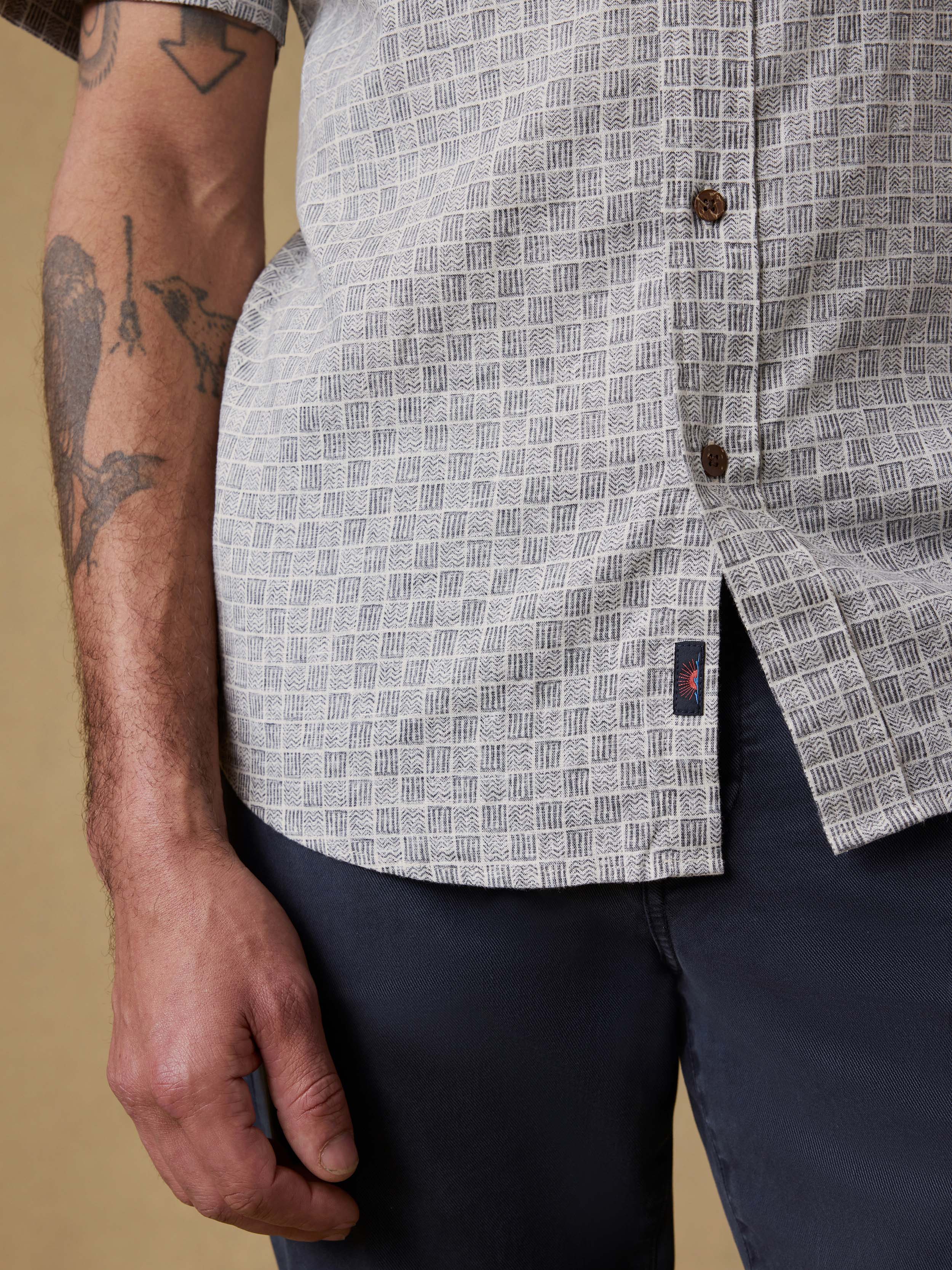 Detail shot of a man wearing a patterned short-sleeve shirt with visible Faherty brand logo and navy pants against a beige background