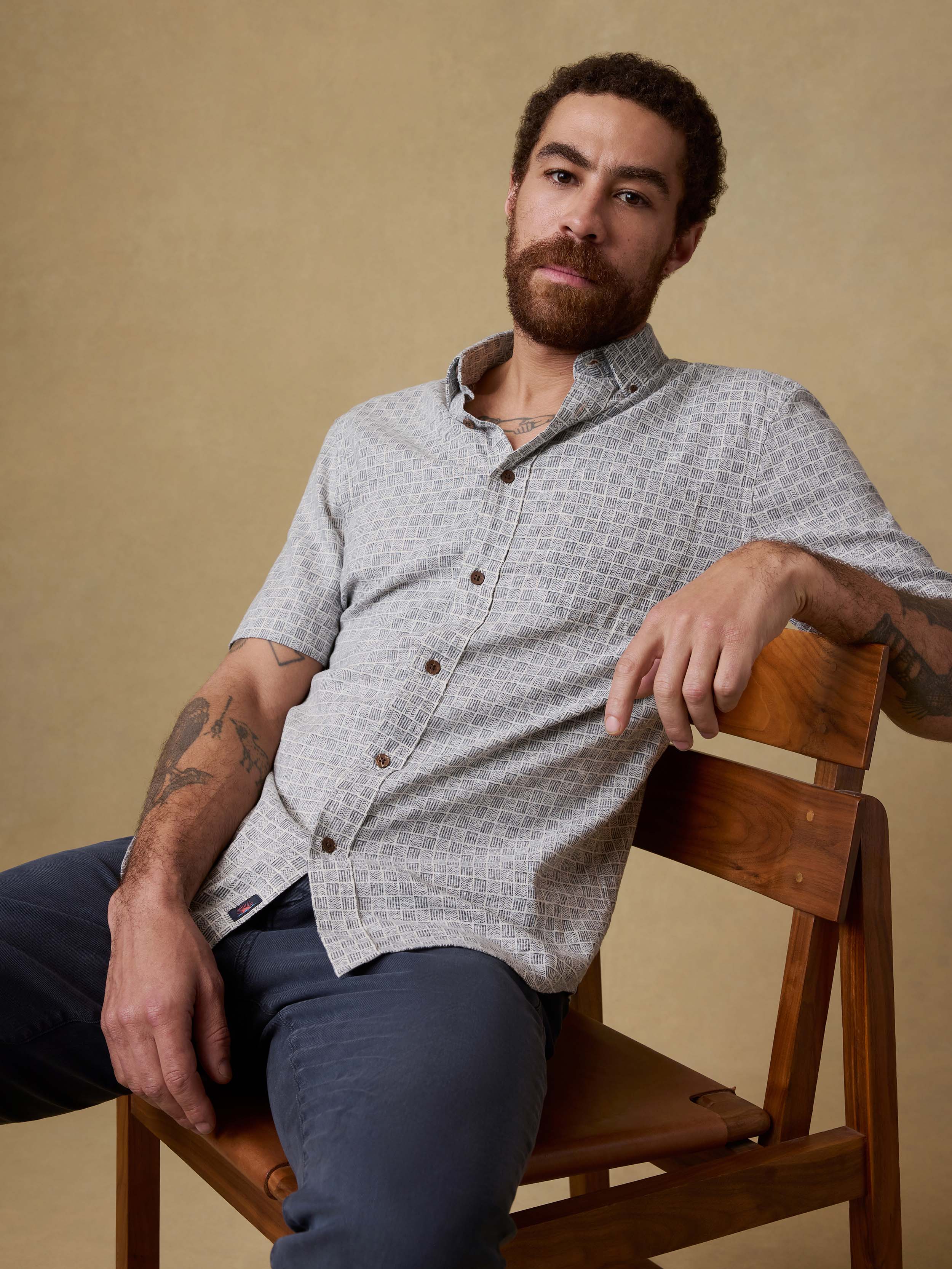 Man sitting on a chair wearing a patterned short-sleeve shirt and navy pants against a beige background