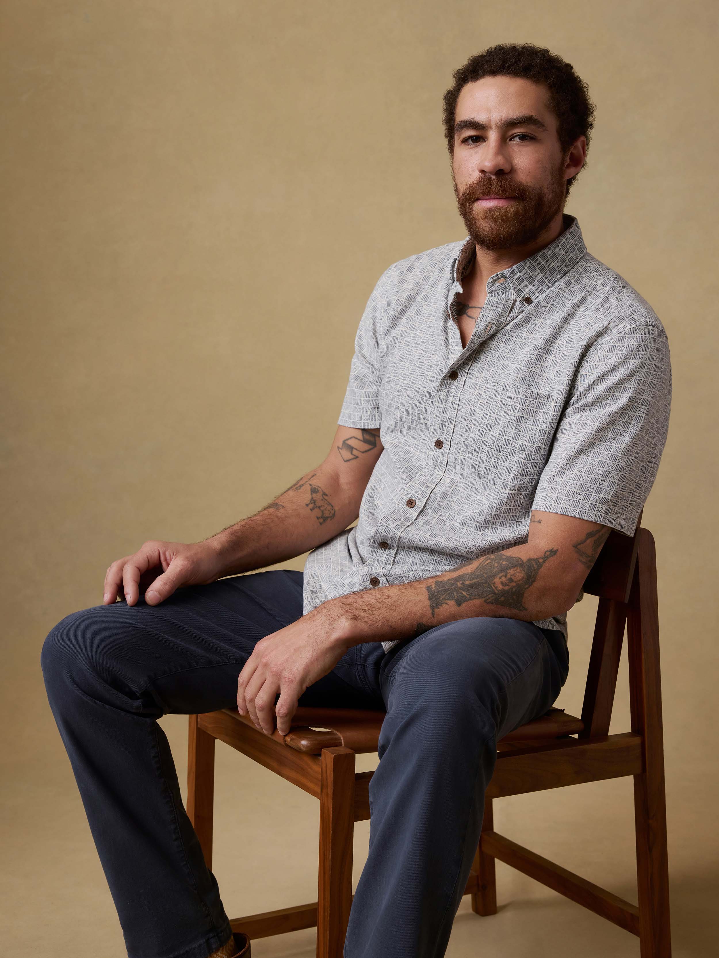 Man sitting on a chair wearing a patterned short-sleeve shirt and navy pants against a beige background